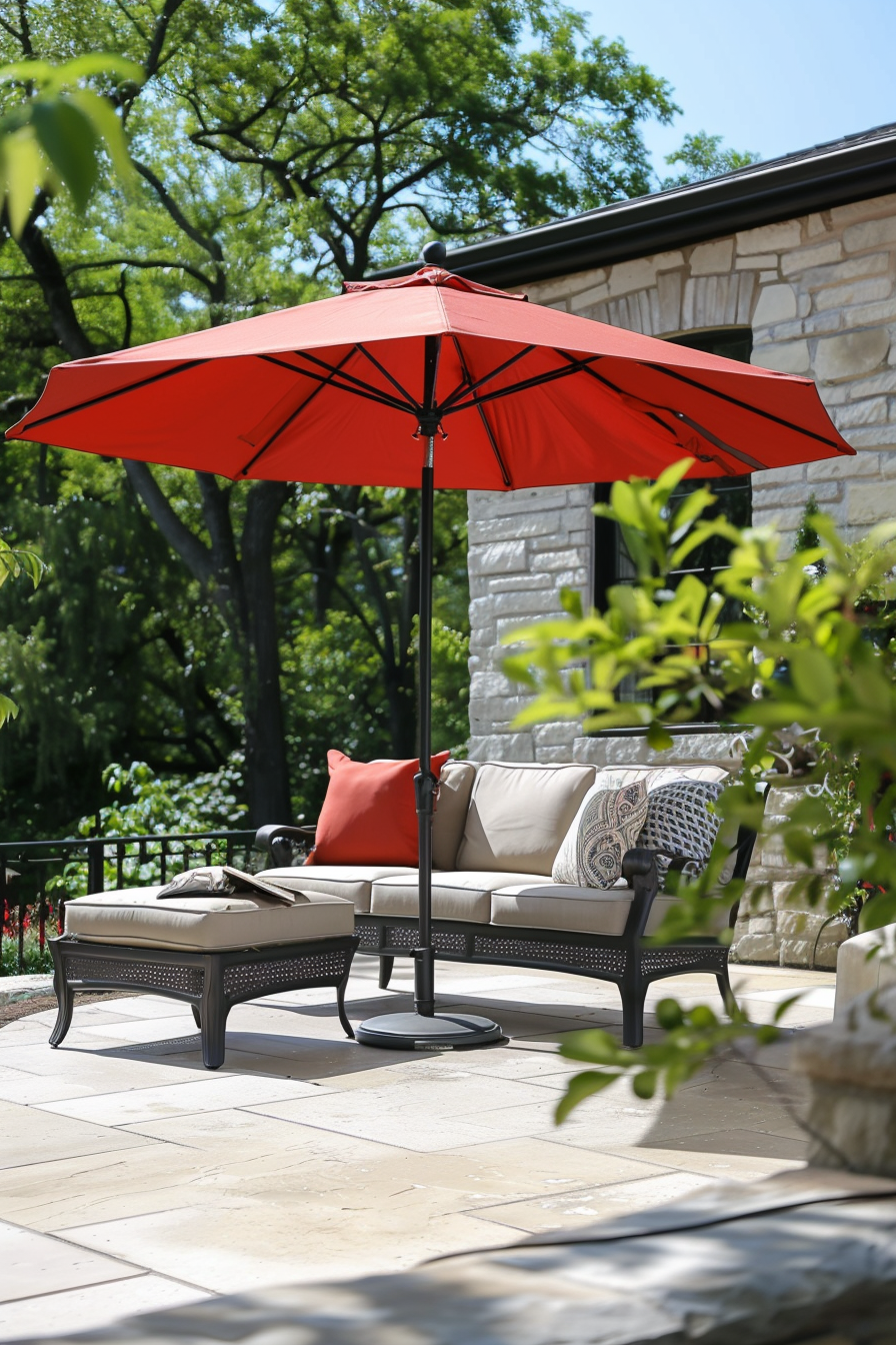 A red patio umbrella providing shade over outdoor furniture on a sunny day, with lush greenery and a stone house in the background.