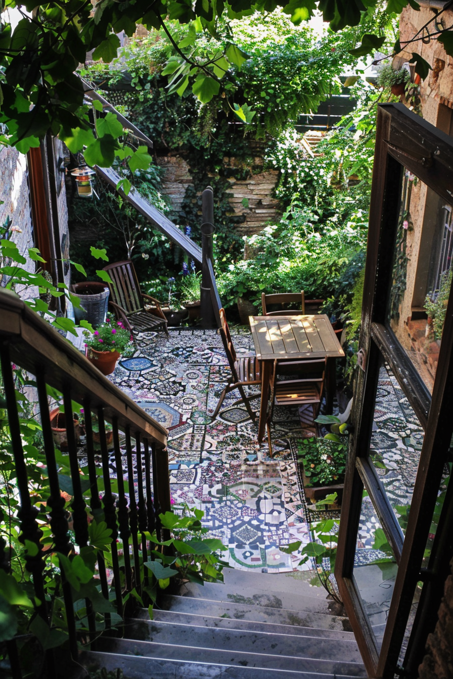 A cozy outdoor patio with patterned tiles, wooden furniture, lush greenery, and a staircase viewed from an open door.
