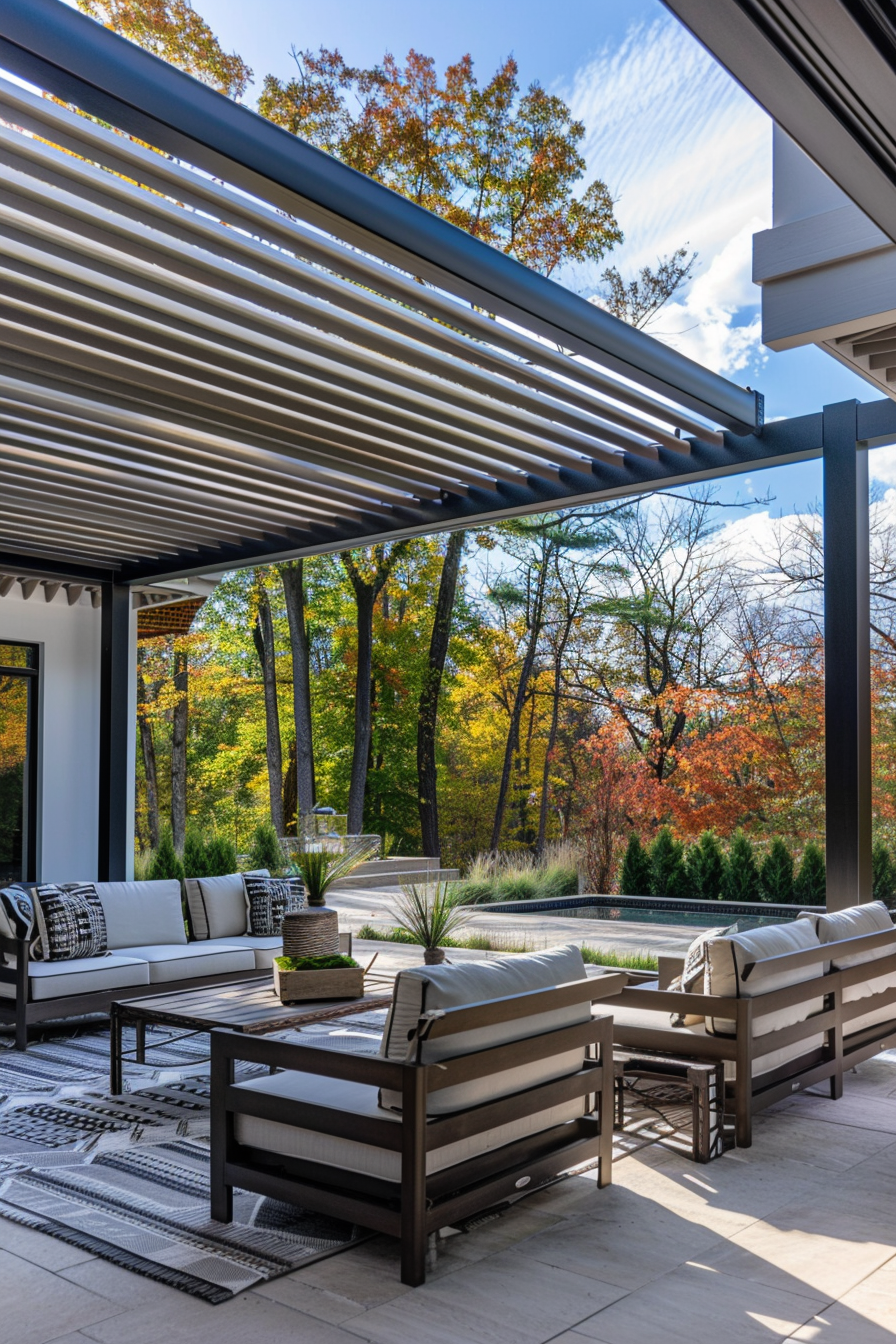 Outdoor patio area with modern furniture under a pergola, overlooking trees with autumn foliage.