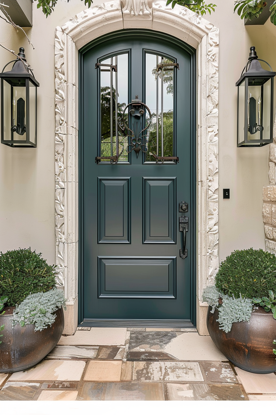Elegant dark teal front door with glass panels, ornate stone trim, flanked by two lanterns and potted plants on a patterned stone porch.