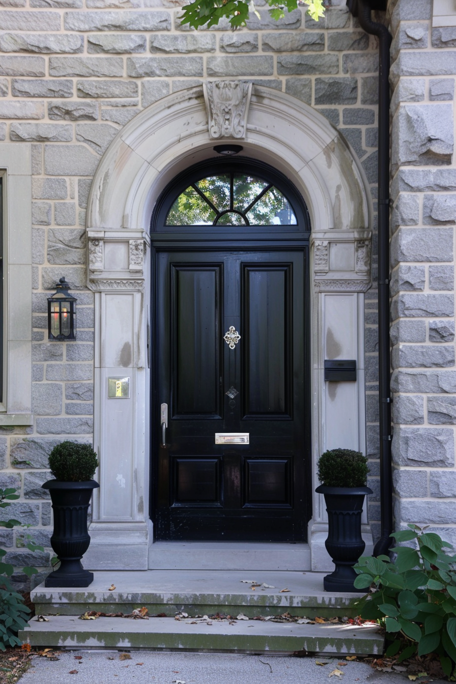 An elegant black arched door with decorative stone surround, flanked by two planters, on a traditional stone building facade.