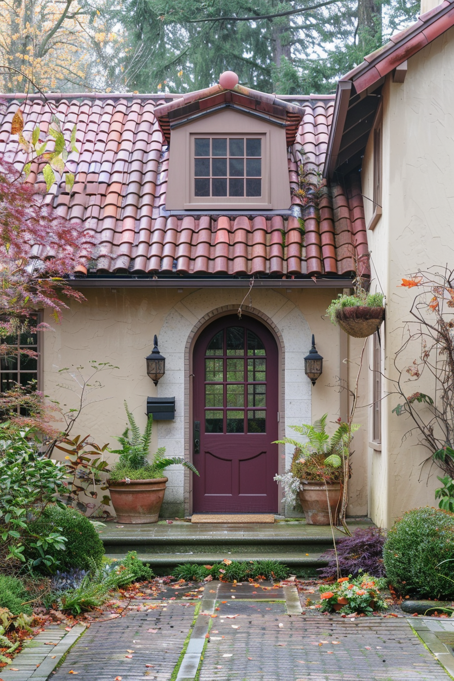 Charming entrance to a house with a burgundy door, surrounded by a lush garden and a tiled pathway with fallen leaves.