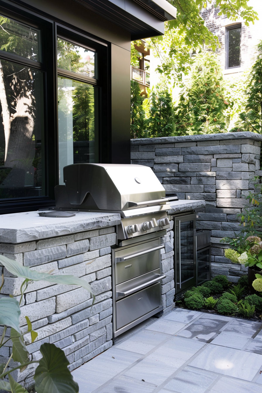 Outdoor grill and refrigerator built into a stone patio kitchenette, with greenery and black trimmed windows in the background.