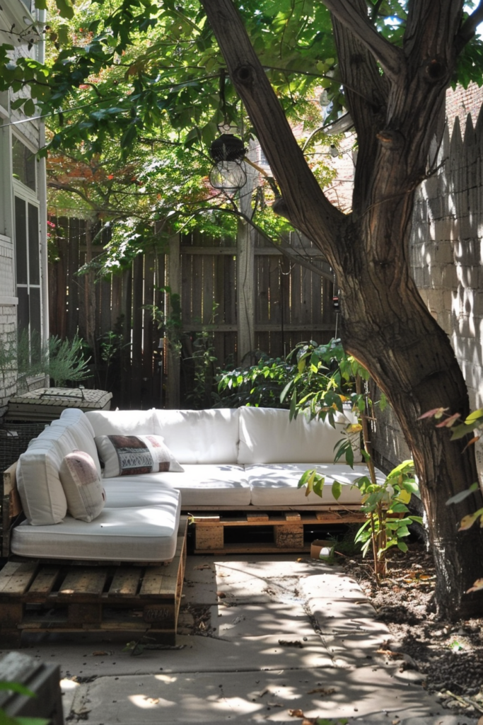 A cozy outdoor seating area with a white couch made from pallets, situated under a tree in a leafy backyard.