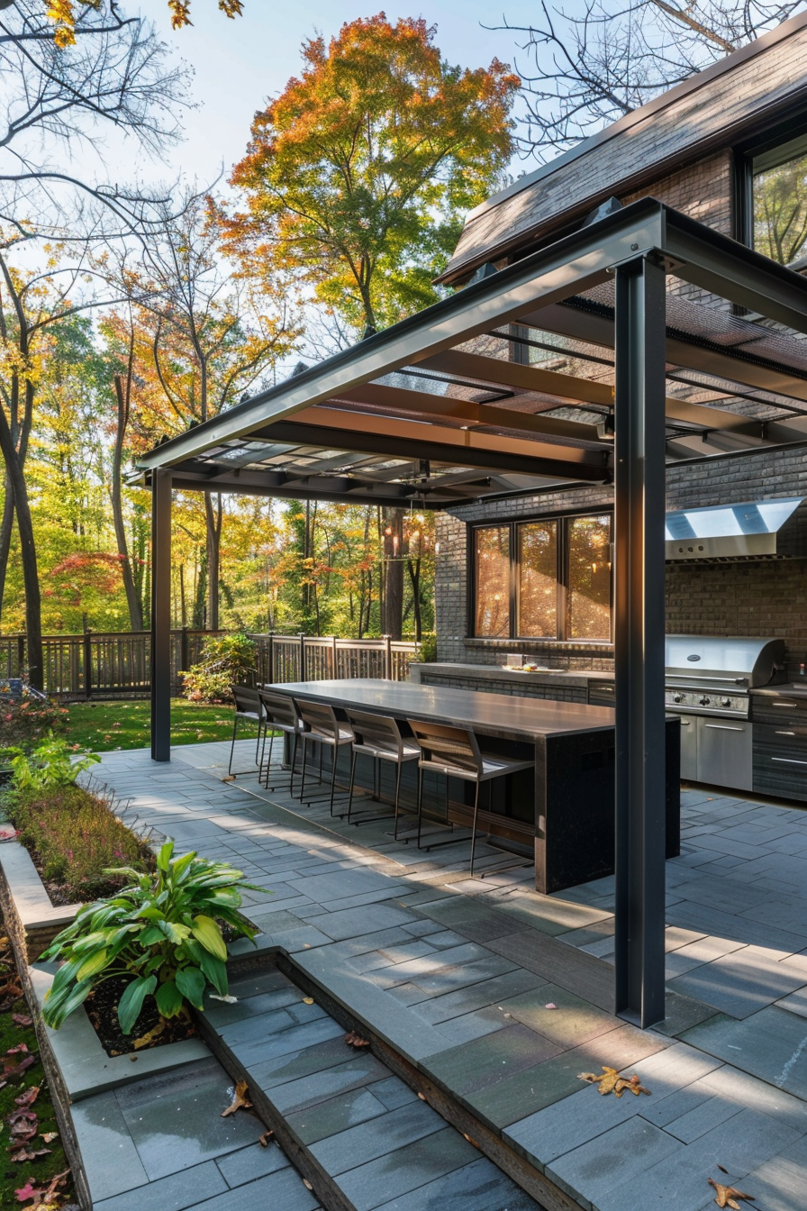 Outdoor kitchen patio with a long dining table, modern grill, under a pergola, surrounded by autumn trees.