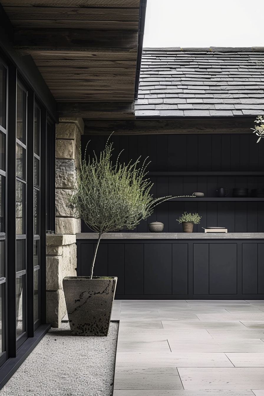 The scene includes a modern outdoor patio area with a weathered stone column supporting a wooden beam. A dark slate roof provides contrast above, while the ground is covered with large, pale tiles that continue inside through glass doors. A large potted plant with thin leaves is prominently displayed in a textured stone container, showing signs of age. In the background is a sleek kitchen with black cabinetry and a counter displaying a smaller plant, books, and minimalist kitchenware, all adhering to a muted, sophisticated color palette. Modern patio with stone planters and a sleek black kitchen interior.