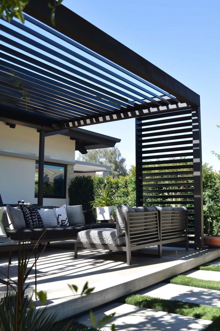 A modern outdoor patio with a pergola, stylish furniture, and decorative cushions, surrounded by lush greenery on a sunny day.