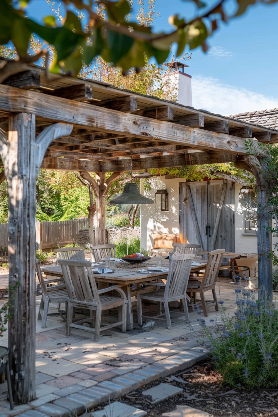 Rustic outdoor dining area with wooden table and chairs under a pergola, greenery around, and a house in the background.