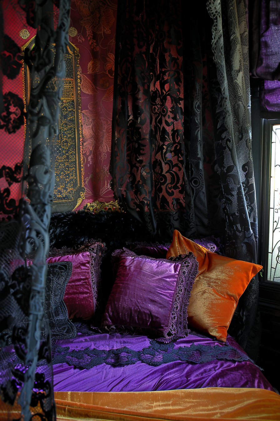 Luxurious bedroom with rich purple and gold bedding, intricate black lace curtains, and ornate wallpaper.
