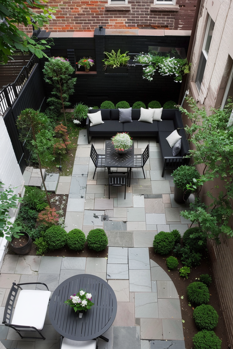 A cozy urban garden patio with black furniture, lush greenery, and patterned stone tiles, viewed from above.