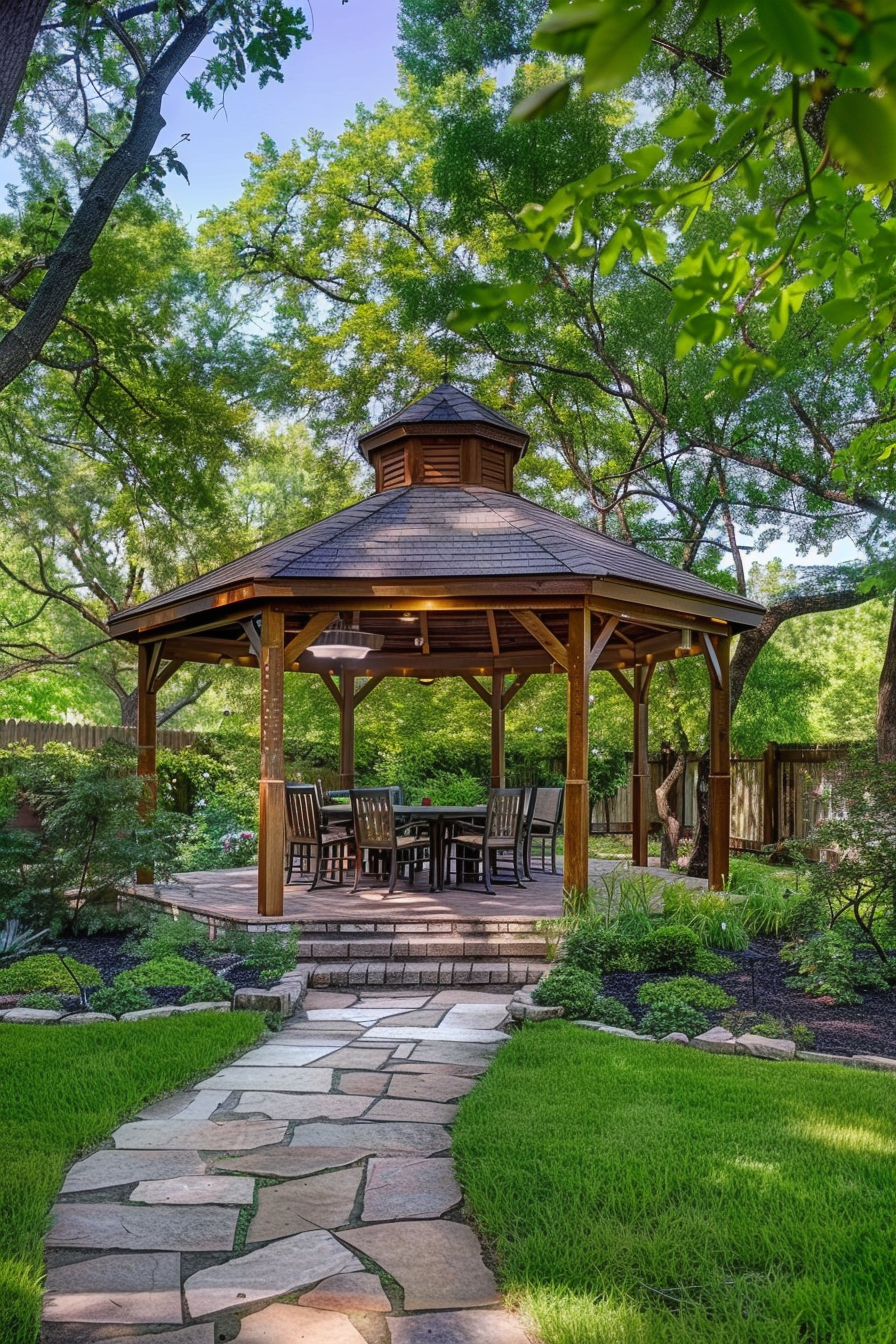A stone pathway leading to a wooden gazebo with a table and chairs set in a lush green garden surrounded by trees.