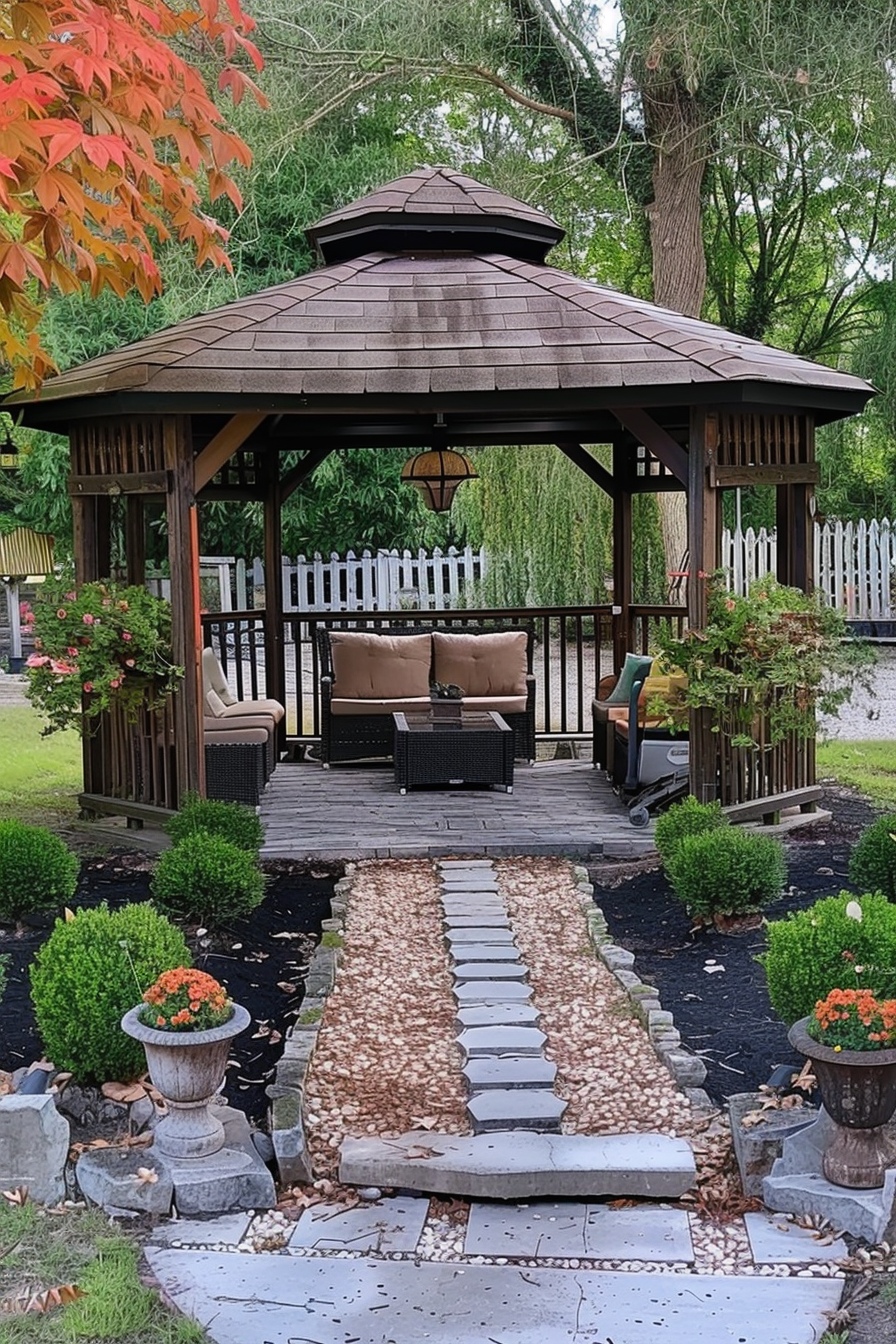 A wooden gazebo with a shingle roof, surrounded by greenery, with a stone pathway leading up to it.