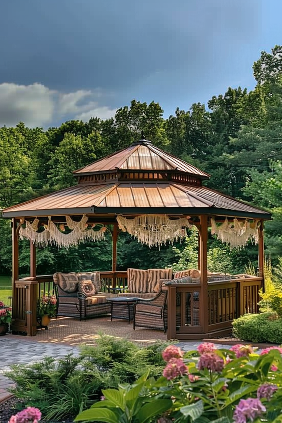 A wooden gazebo with a metal roof, surrounded by lush greenery and flowers, featuring cozy seating and decorative hanging lights.