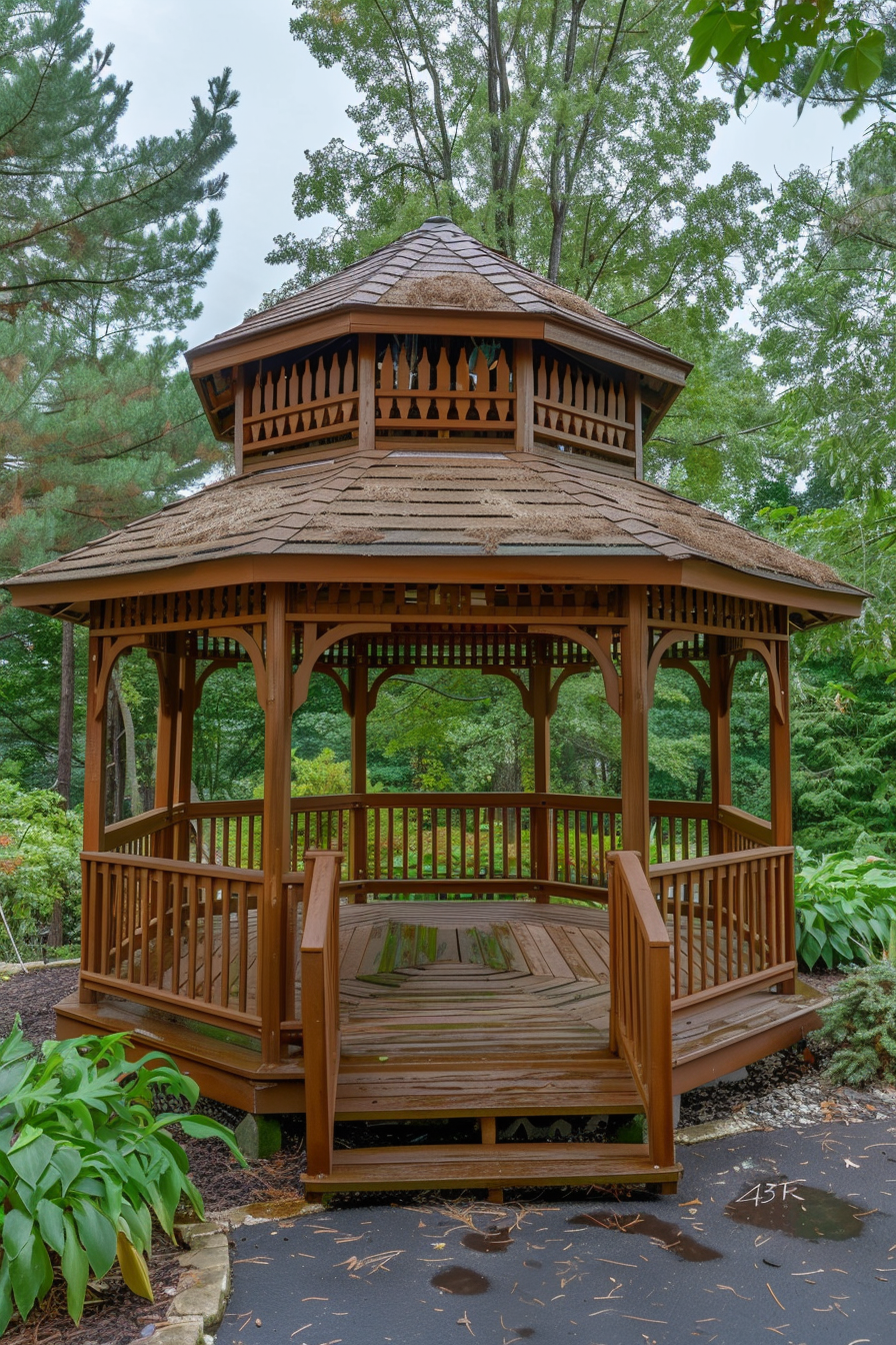 Wooden gazebo with a shingled roof in a lush garden surrounded by tall trees and greenery.