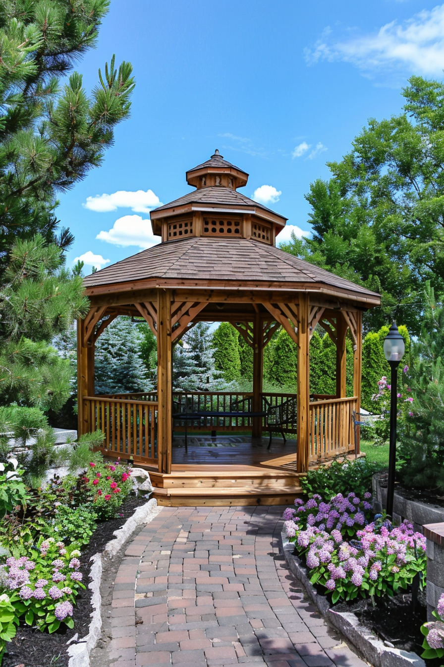 A wooden gazebo with a shingled roof on a brick pathway, surrounded by greenery and blooming purple flowers under a clear blue sky.