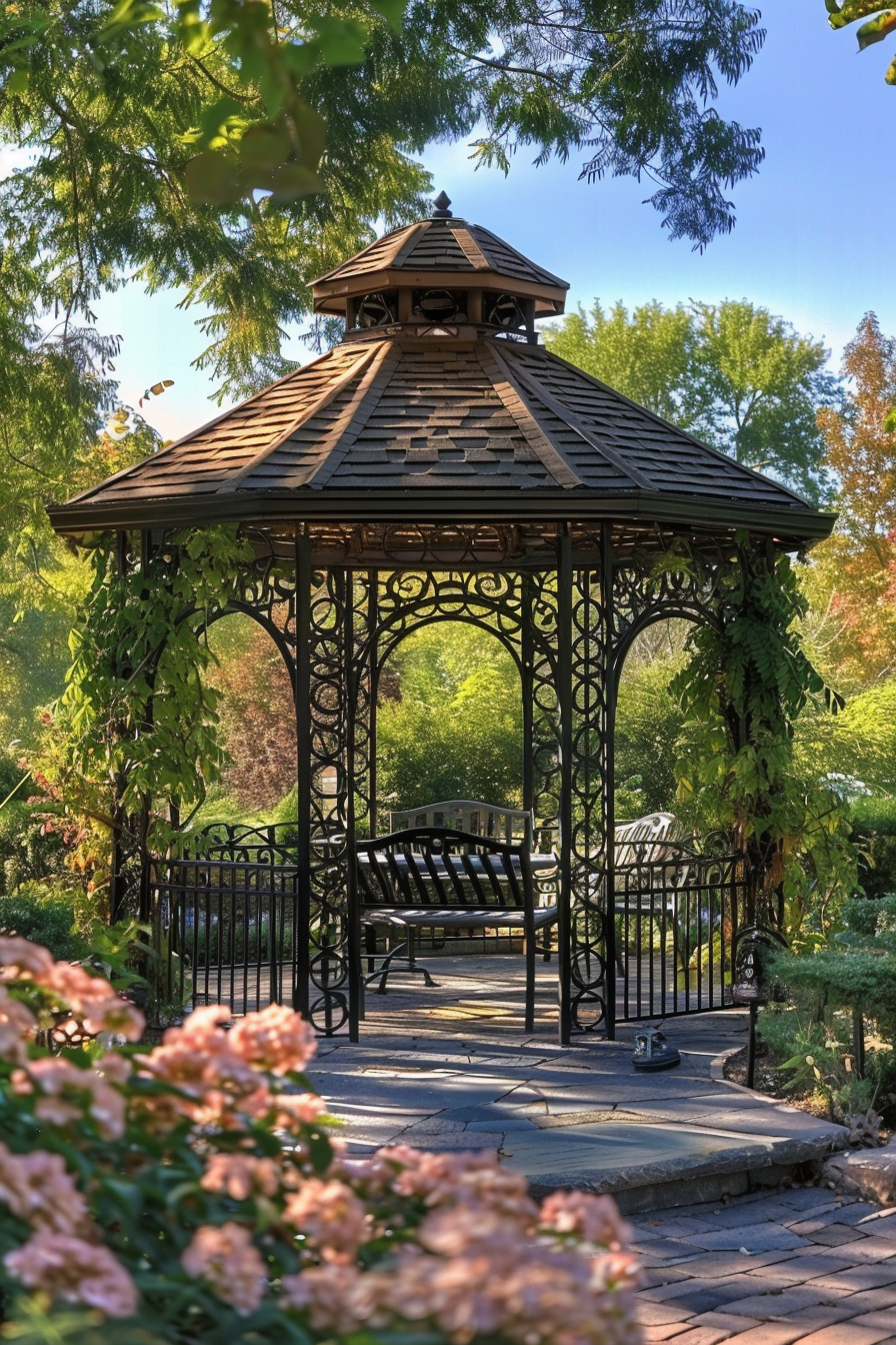 Alt text: An ornate black metal gazebo with benches, surrounded by blooming flowers and lush greenery in a sunlit garden setting.