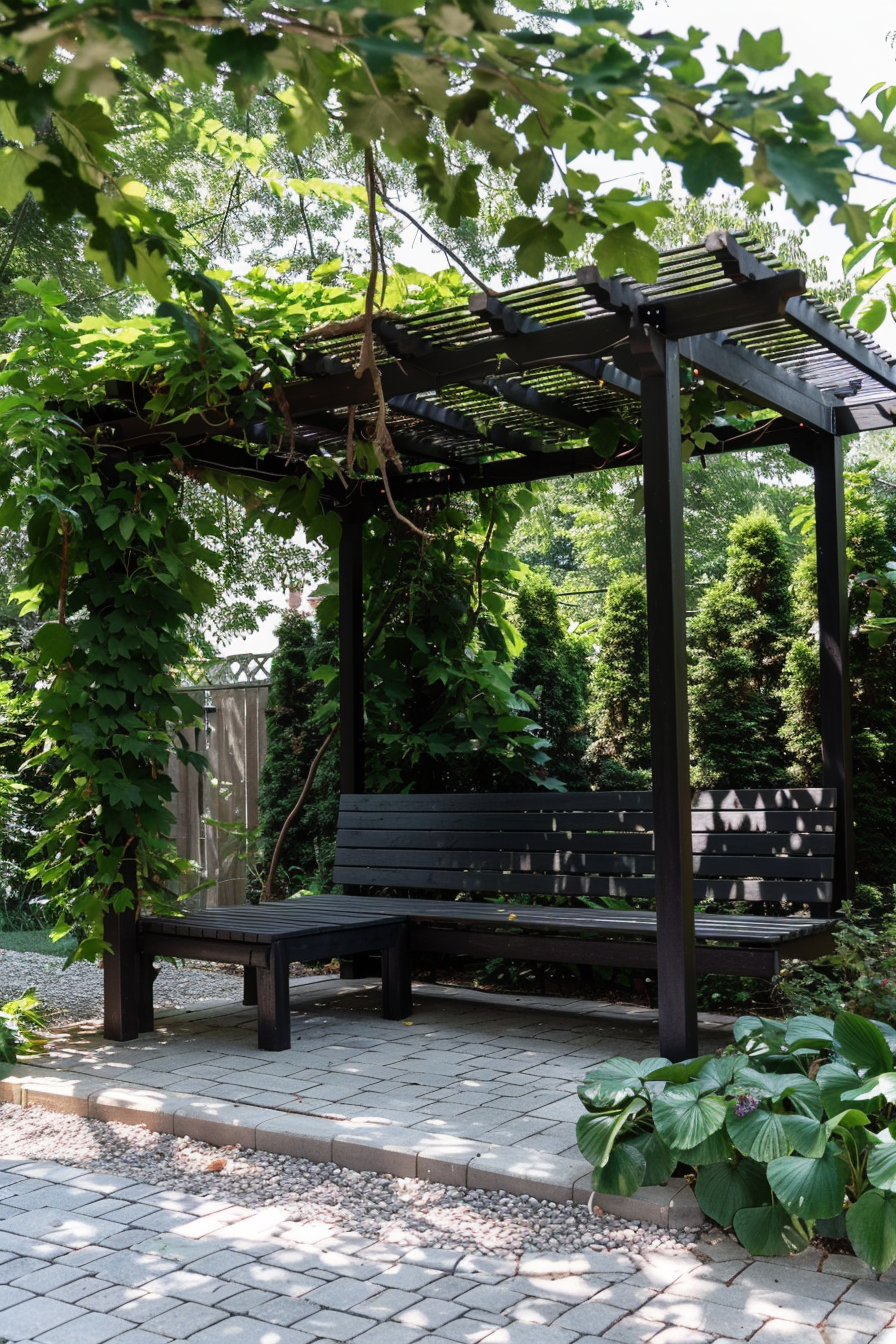 Wooden garden bench under a pergola draped with lush green vines, surrounded by leafy plants and paved ground.