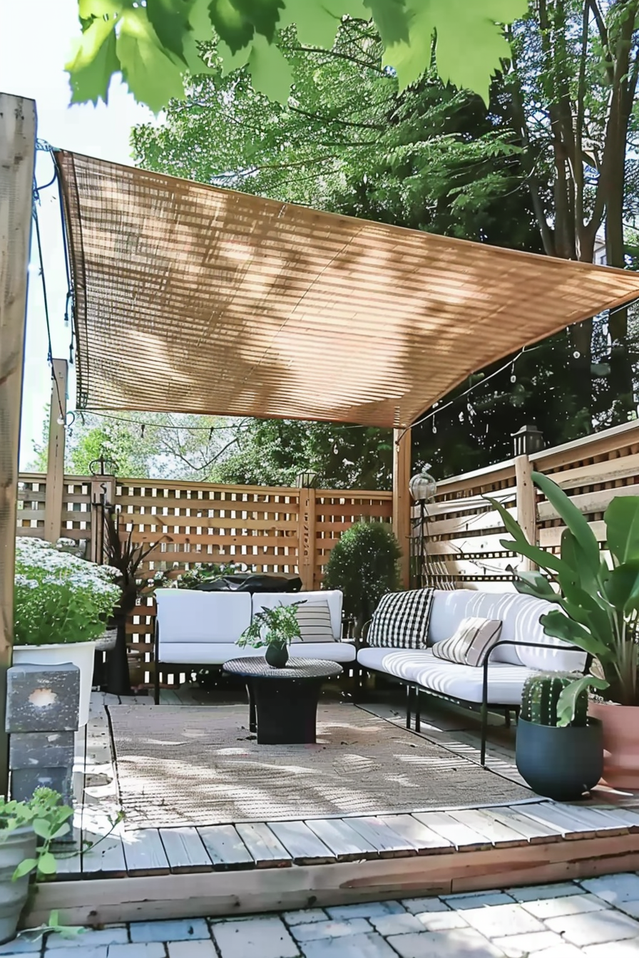 A cozy outdoor patio with a sectional couch, a coffee table, plants, and a sunshade, surrounded by a wooden fence.