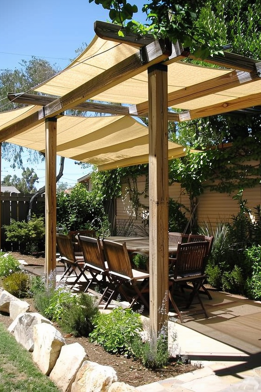 A wooden pergola with beige fabric shades over a patio with chairs, surrounded by greenery and large rocks.