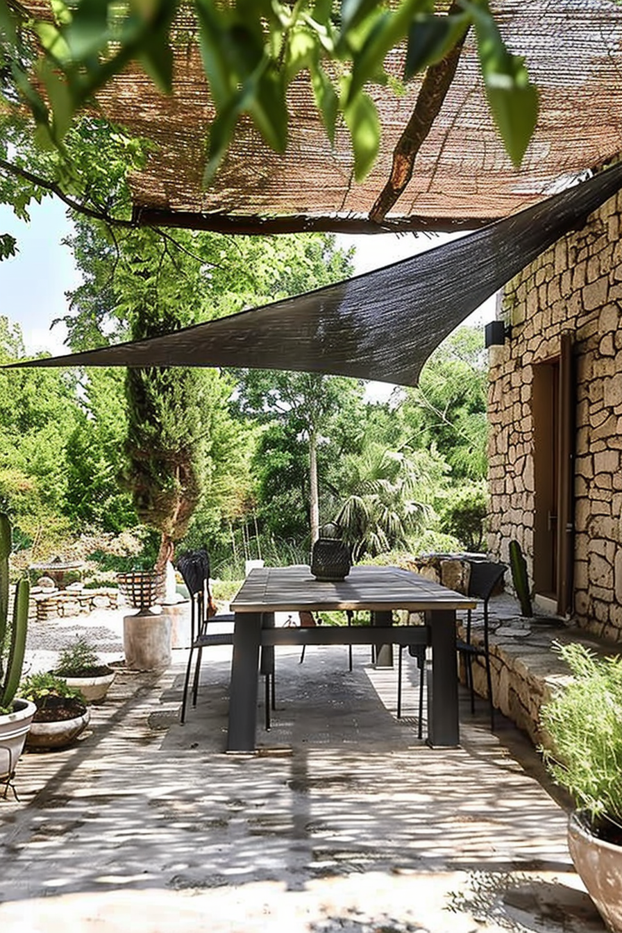 ALT: A serene patio space with a large dining table, shaded by a canopy and surrounded by greenery, attached to a stone-built house.