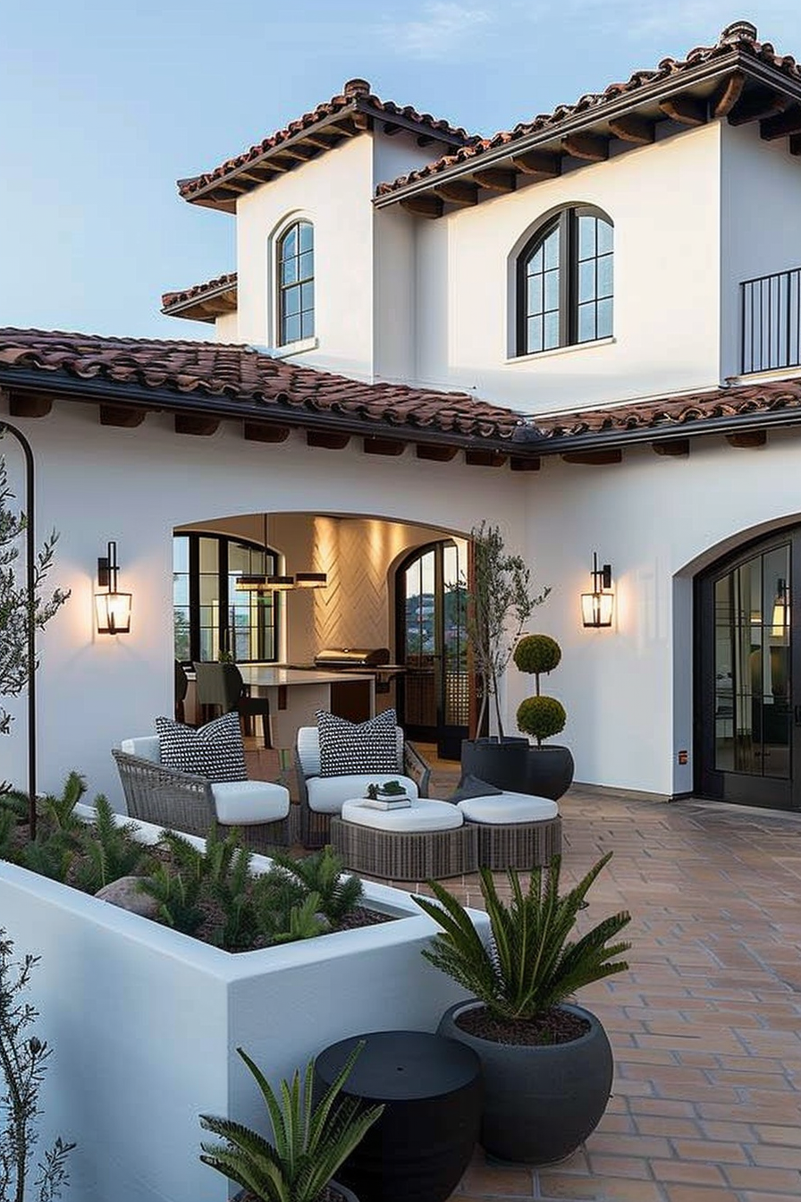 Elegant outdoor patio with seating, potted plants, and wall-mounted lanterns, adjacent to a white Mediterranean-style house at dusk.