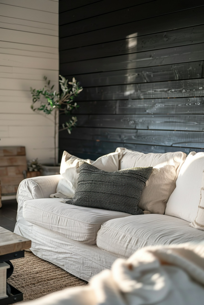 A cozy living room corner with a white couch adorned with textured cushions, against a contrasting black wooden wall bathed in sunlight.
