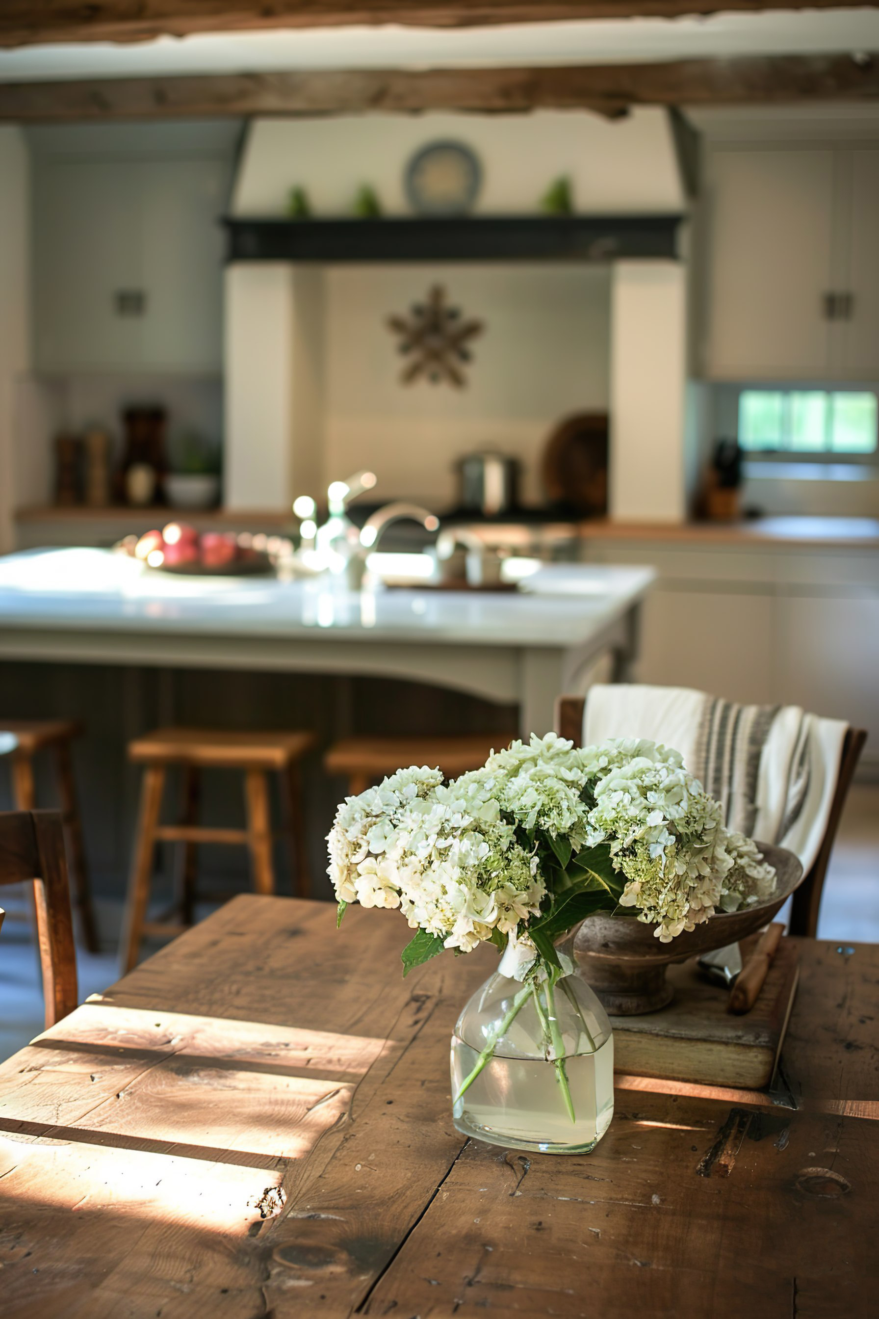 A cozy, sunlit kitchen with wooden beams, a bouquet of white hydrangeas on a rustic table, and a blurred background.