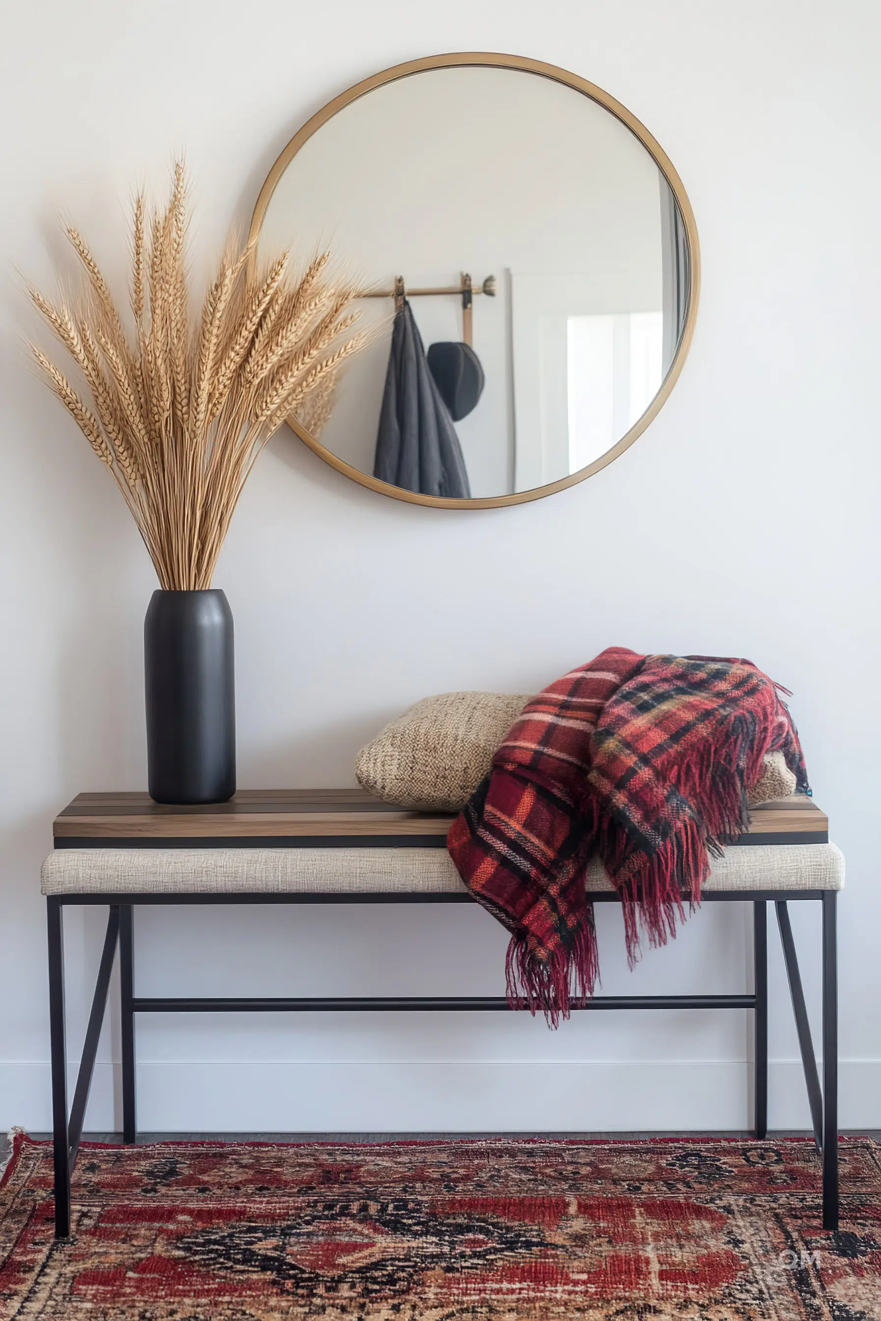A modern entryway with a round mirror, a decorative vase with wheat stalks, a bench with a cushion and throw blanket, and a patterned rug.