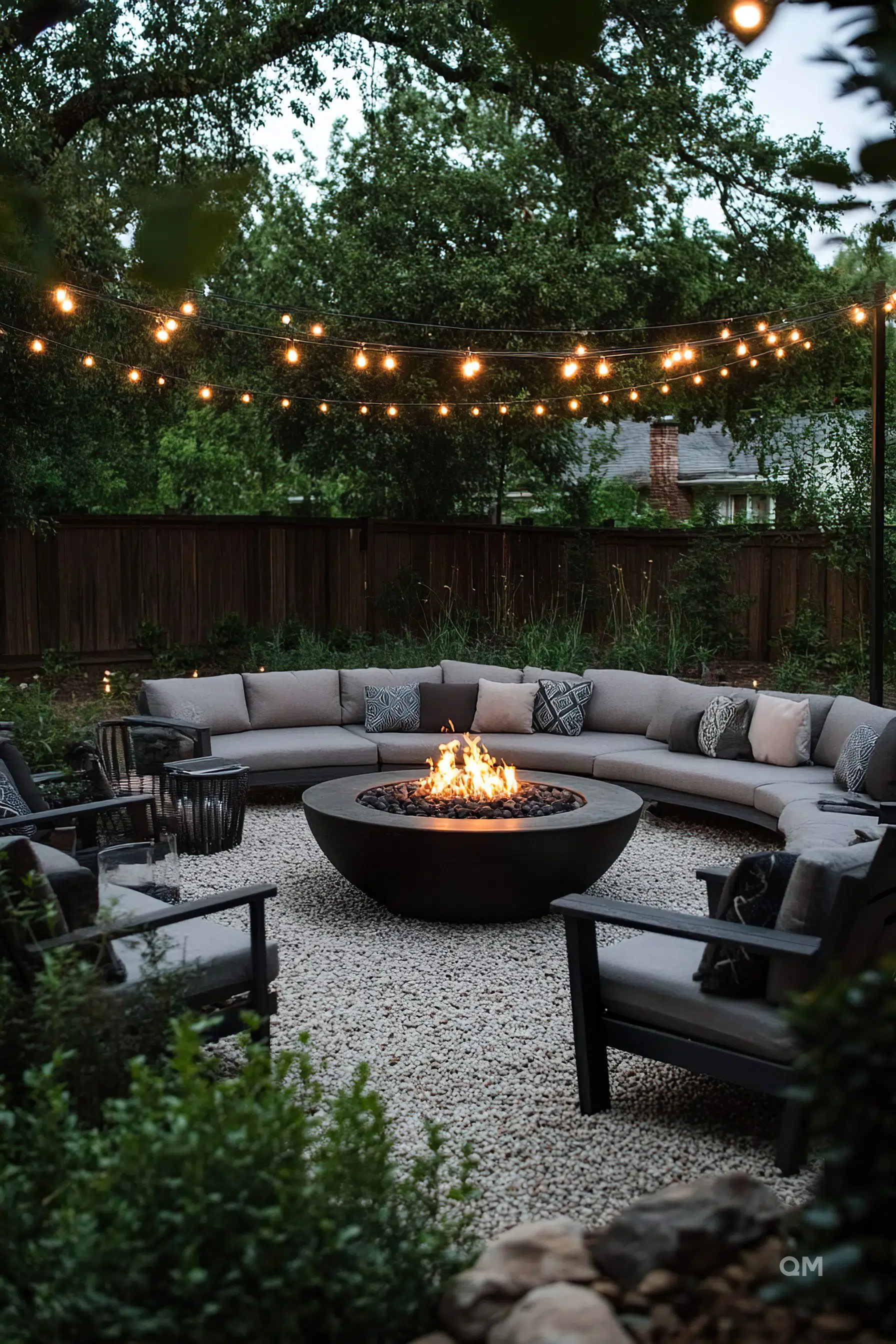 A cozy outdoor seating area with a sectional sofa around a fire pit, string lights above, and lush greenery.