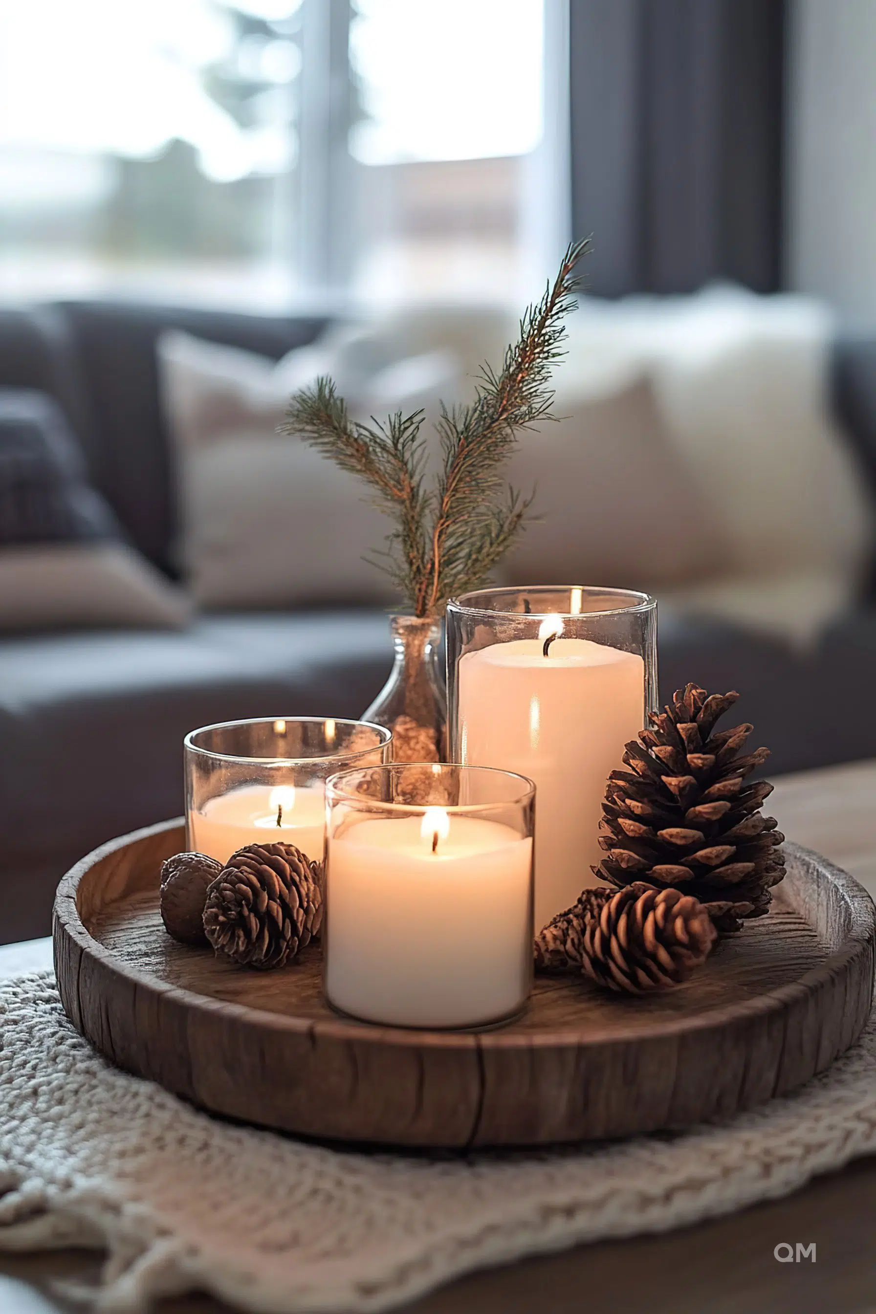 Three lit candles on a wooden tray with pinecones and a sprig of pine, creating a cozy atmosphere in a living room setting.