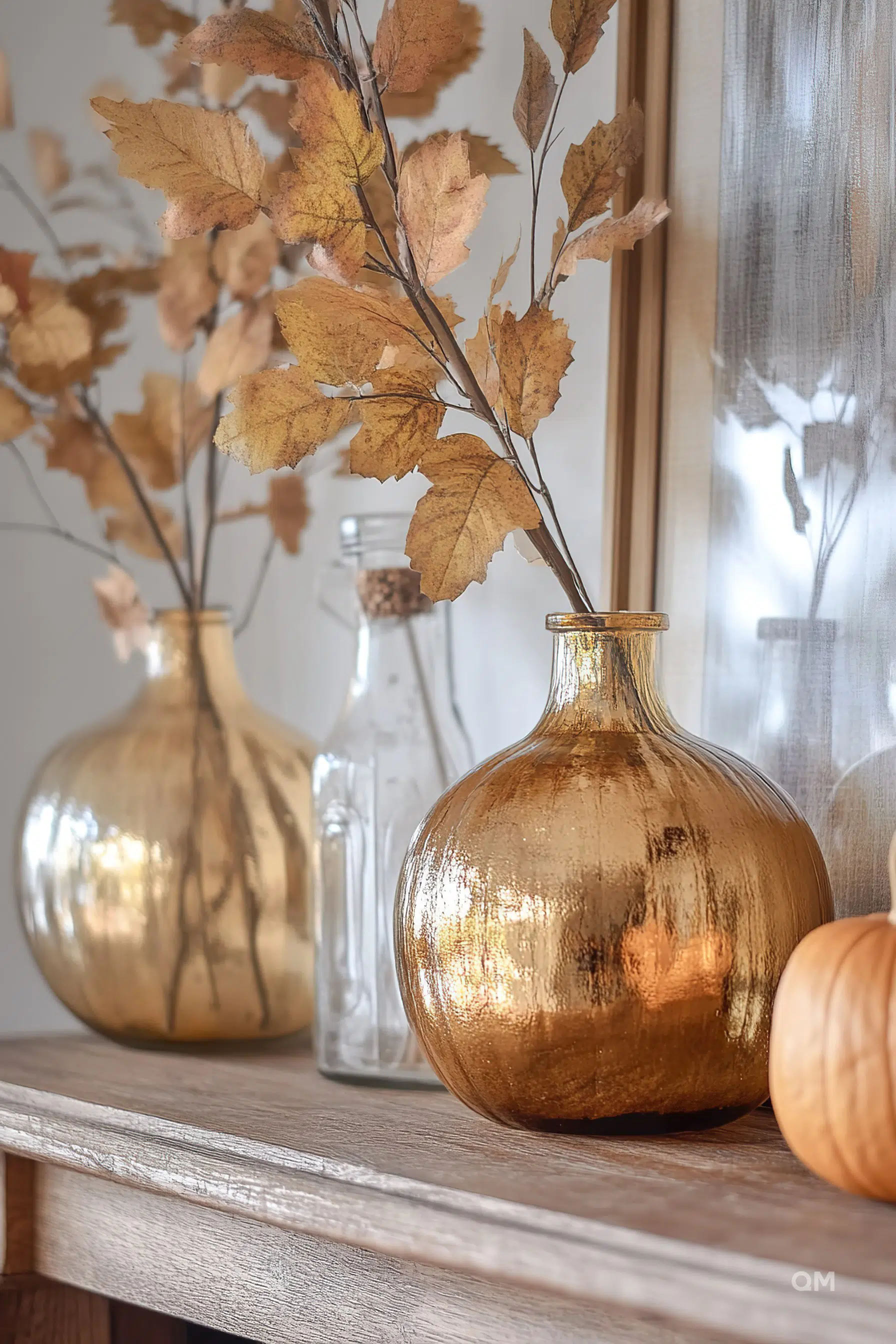 A golden vase with dried leaves on a wooden shelf, accompanied by a clear glass bottle and a mini pumpkin. A cozy, autumnal ambiance.