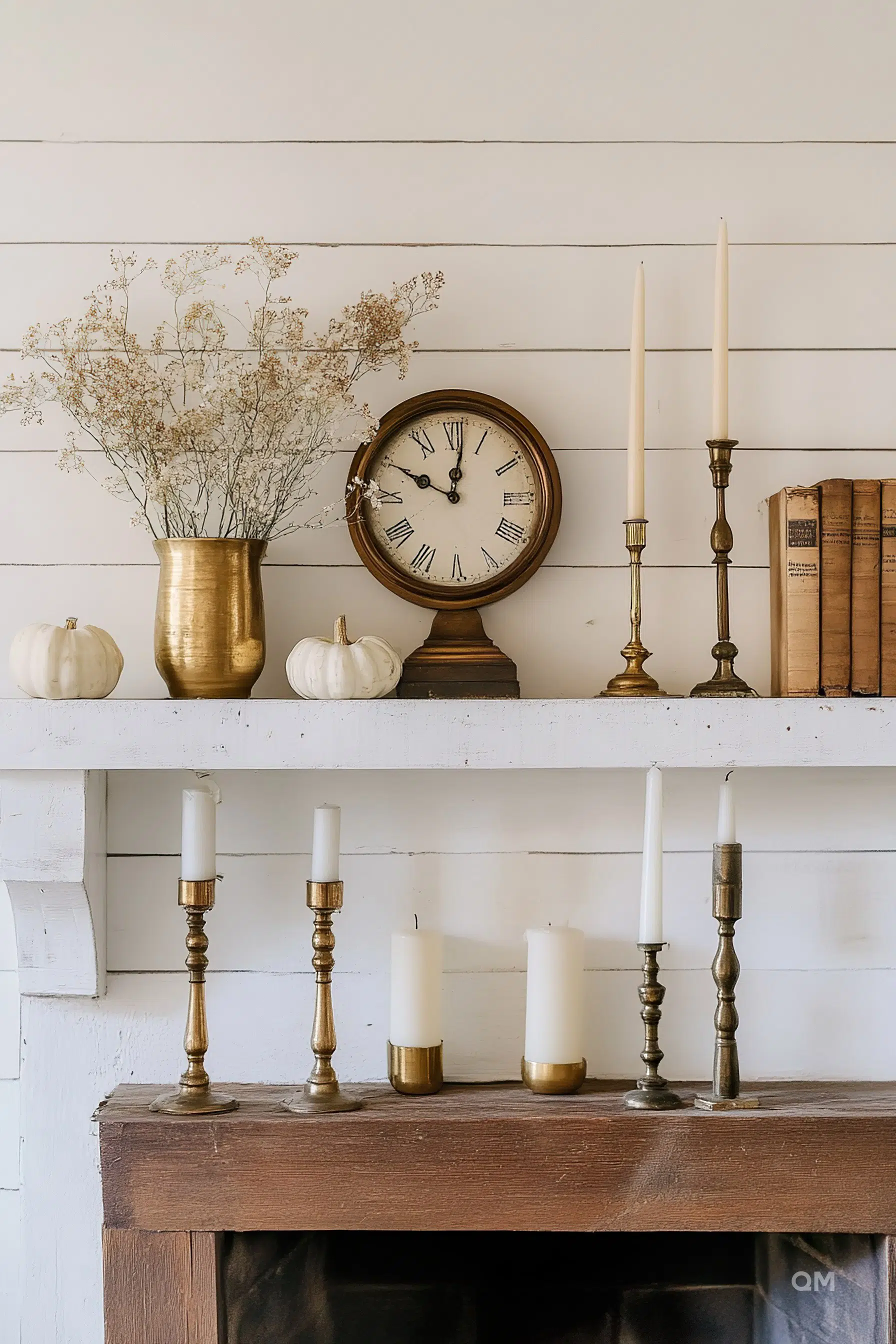 A rustic mantelpiece with an antique clock, brass candlesticks, white pumpkins, and a vase of dried flowers, above a wood fireplace.