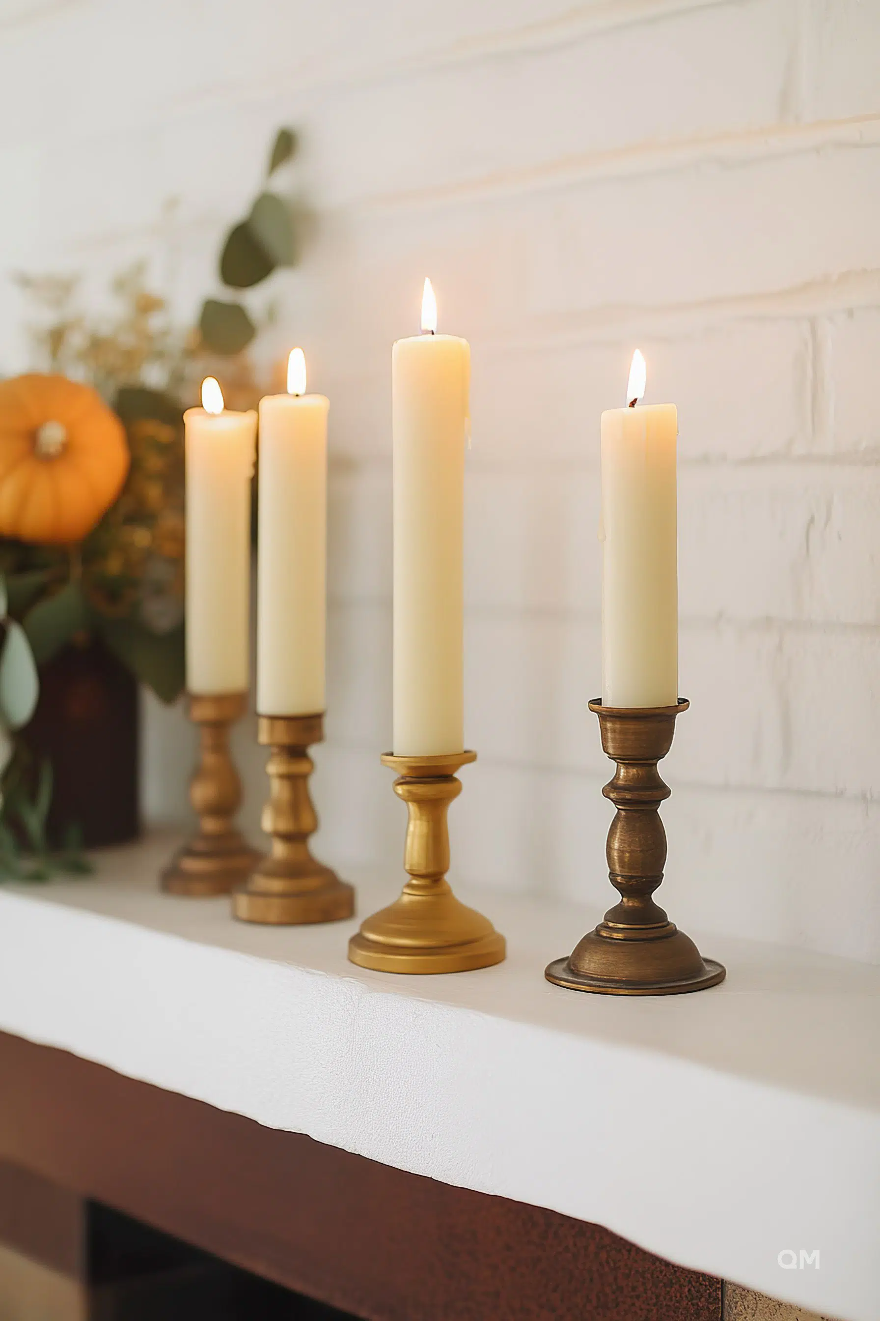 ALT: Four lit ivory candles on brass candlesticks, arranged on a white mantelpiece with a pumpkin and greenery in the background.