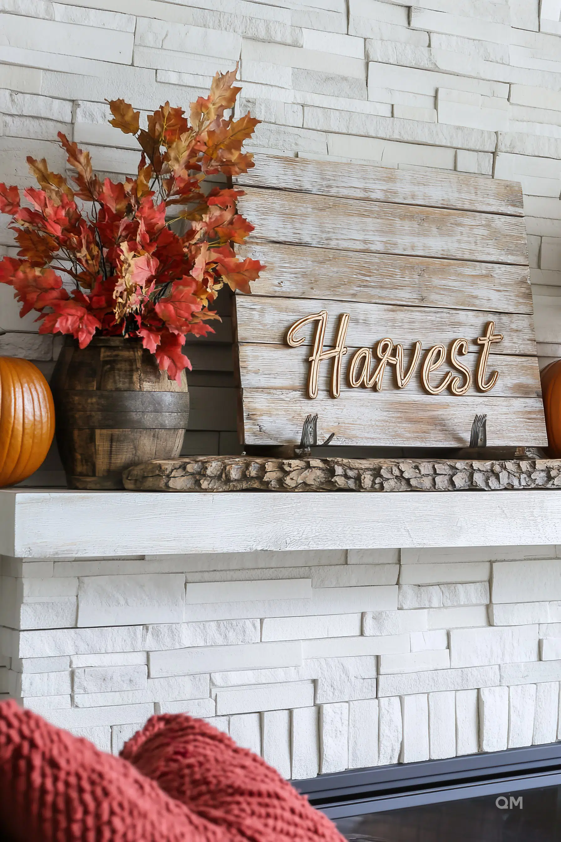 Autumn-themed mantel decor with a wooden "Harvest" sign, fall leaves in a rustic vase, pumpkins, and a cozy knitted throw in the foreground.
