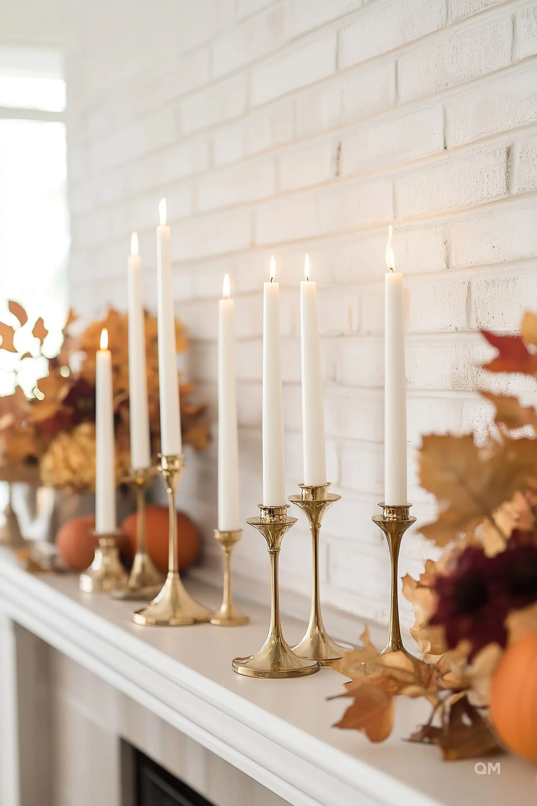 ALT text: "Lit white candles on golden stands set on a mantelpiece with autumn decorations, including leaves and pumpkins, against a brick wall."