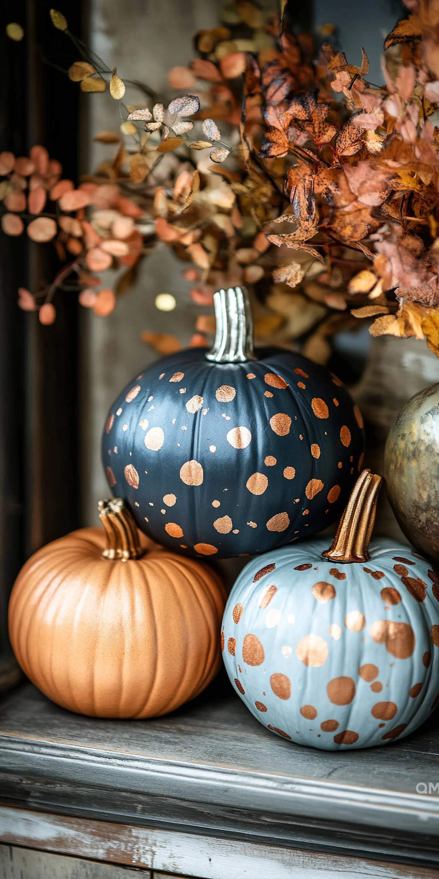 Decorative painted pumpkins in orange, blue with polka dots, and metallic tones alongside autumn leaves on a wooden shelf.