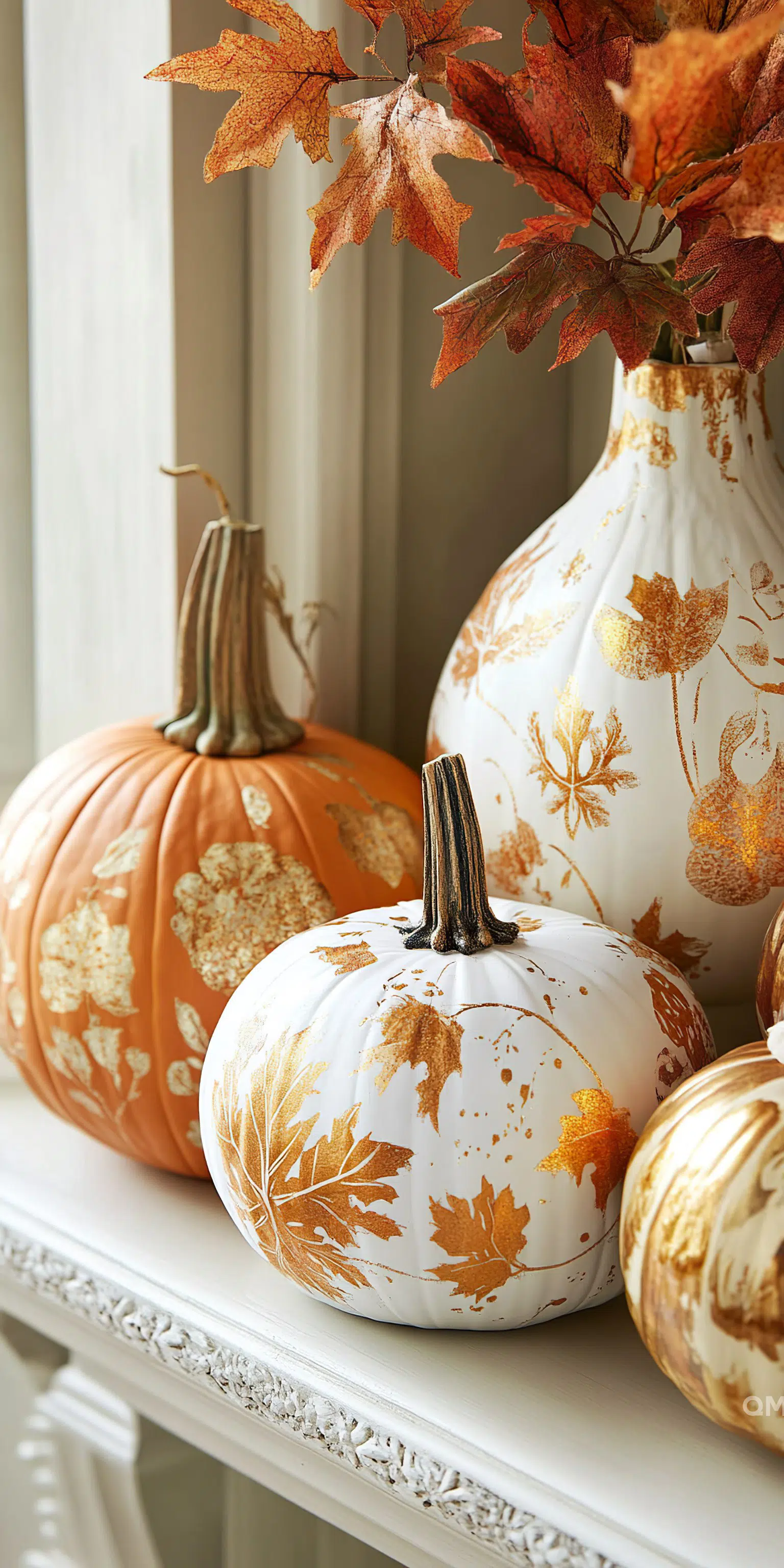 Decorative autumn-themed pumpkins in orange, white, and gold with leaf patterns on a white shelf, accompanied by a vase with fall leaves.