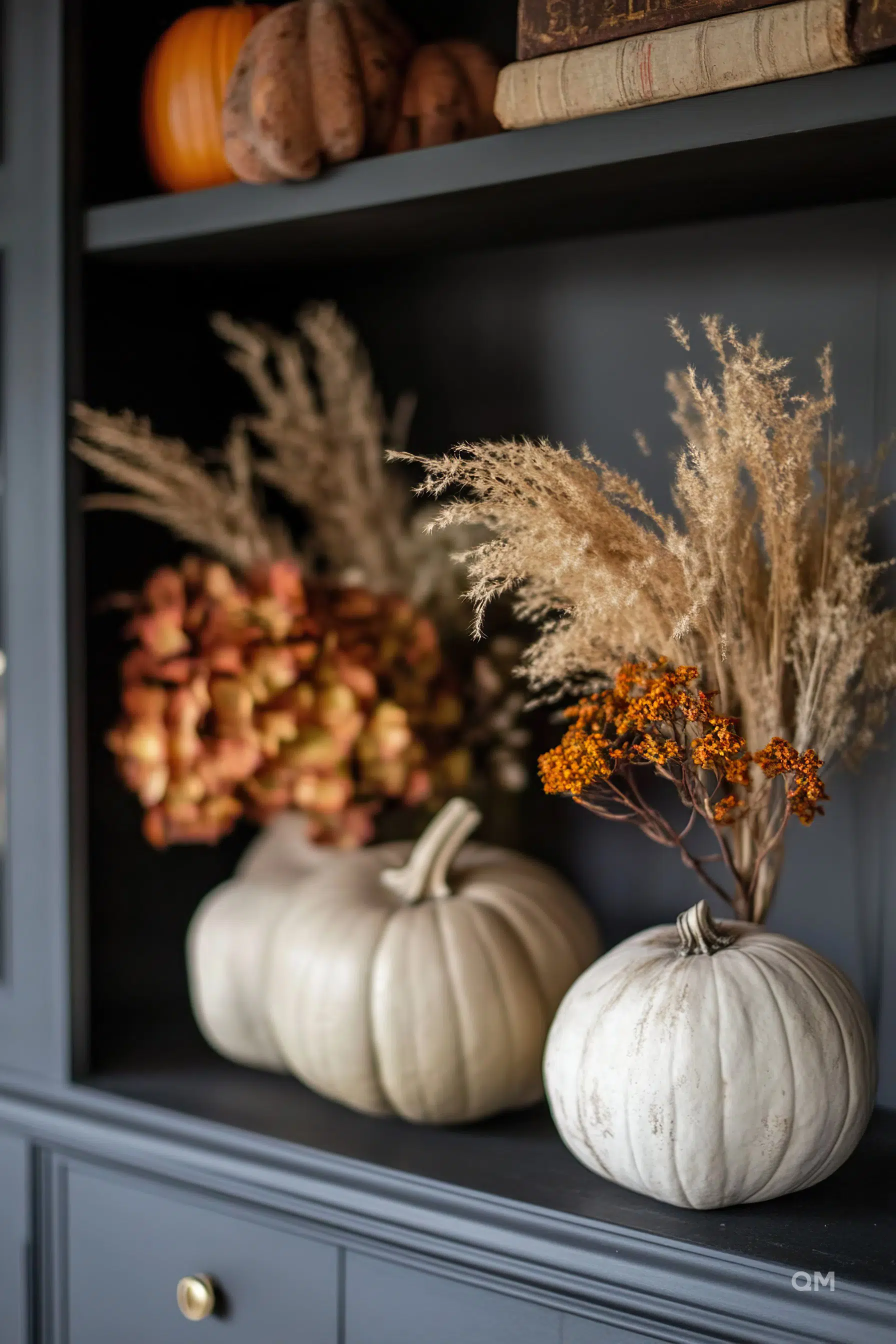 ALT text: Two white decorative pumpkins on a dark shelf alongside dried fall foliage with blurred books and orange pumpkin in the background.