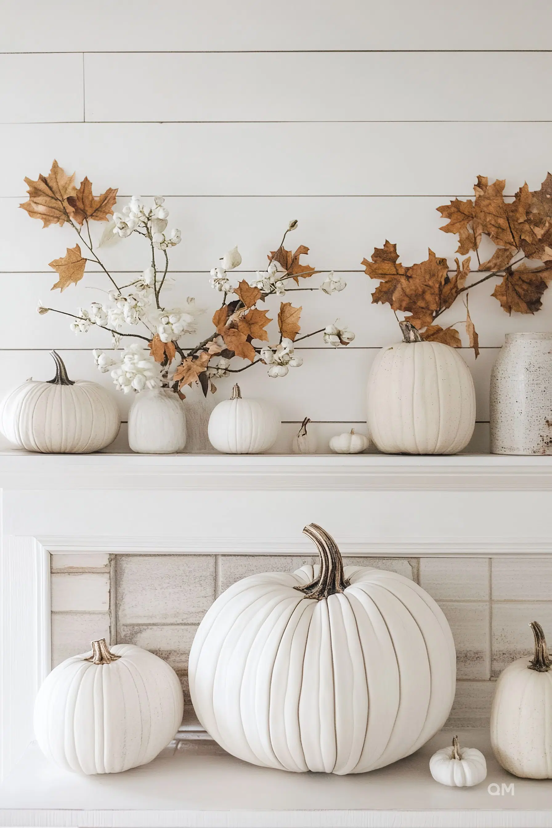 A collection of white pumpkins of various sizes displayed on and in front of a mantelpiece, accented with fall leaves and cotton branches.