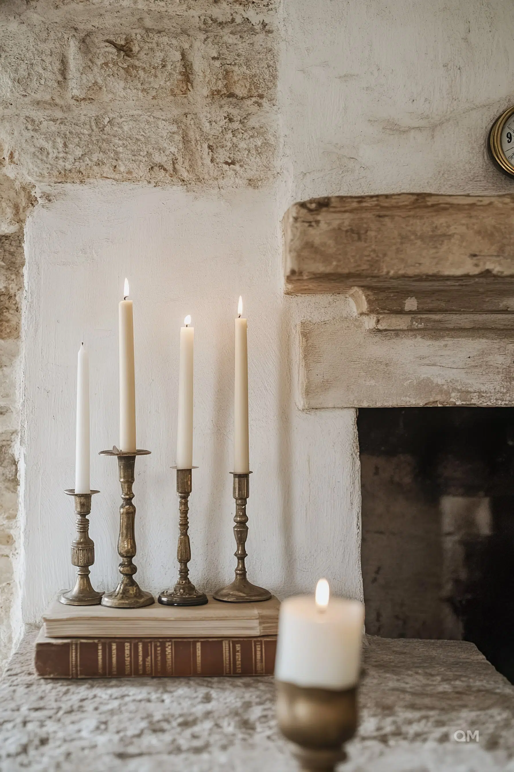 Five lit candles on brass candlesticks, placed on books by a rustic stone fireplace.