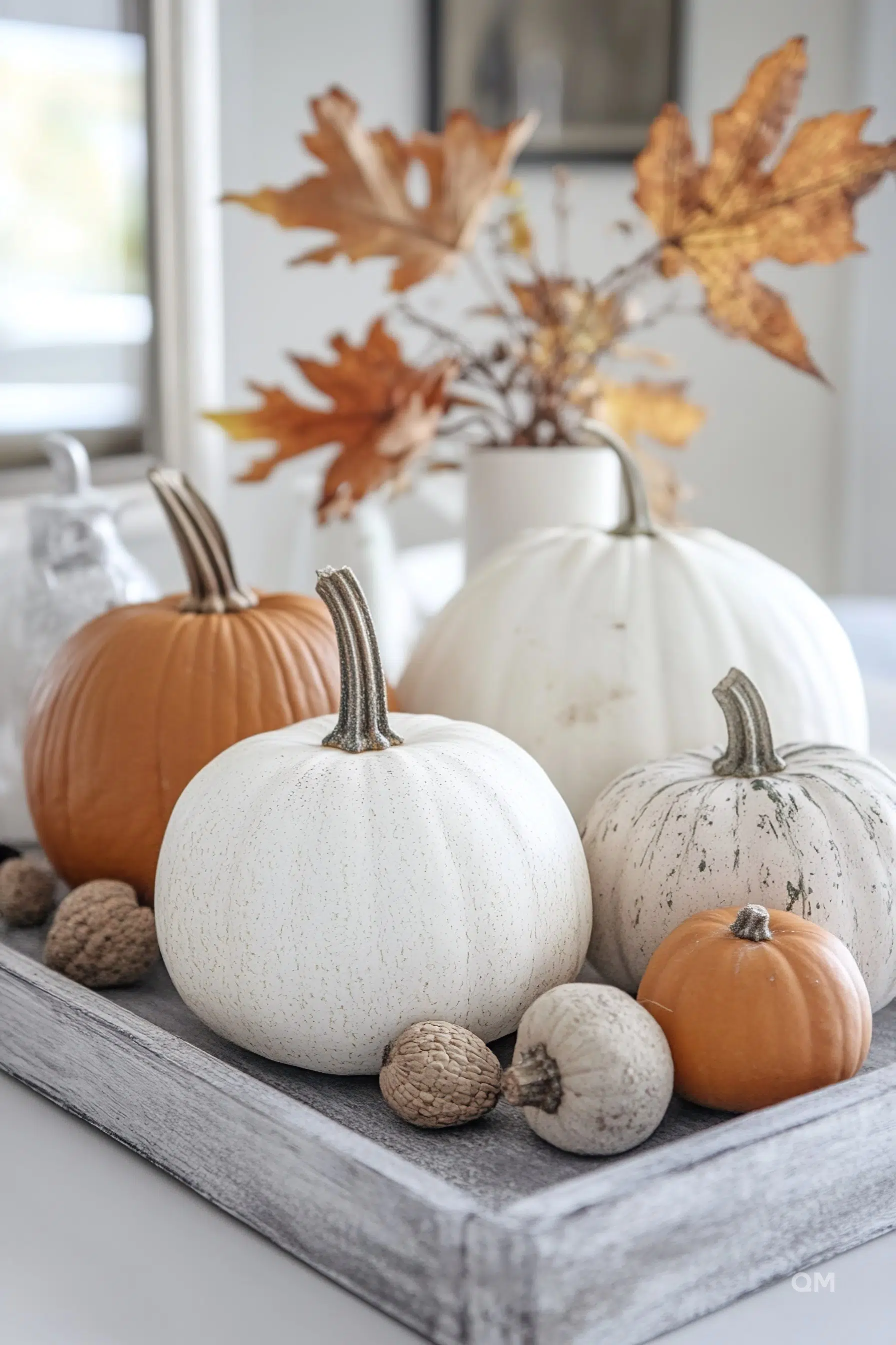 A decorative autumnal display with white, orange, and speckled pumpkins arranged on a wooden tray with acorns and dried leaves in the background.
