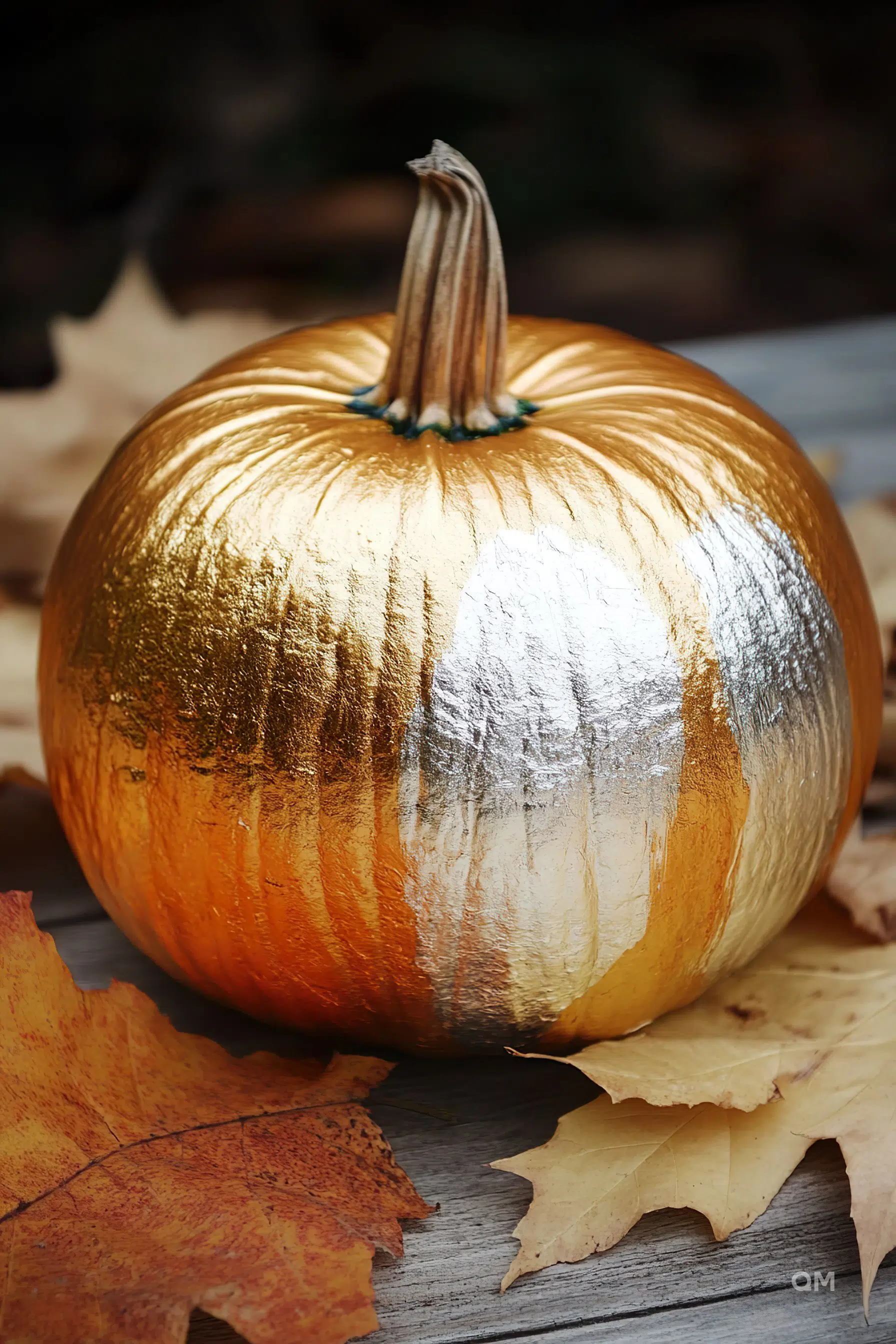 A pumpkin painted with metallic gold and silver sitting amongst autumn leaves on a wooden surface.