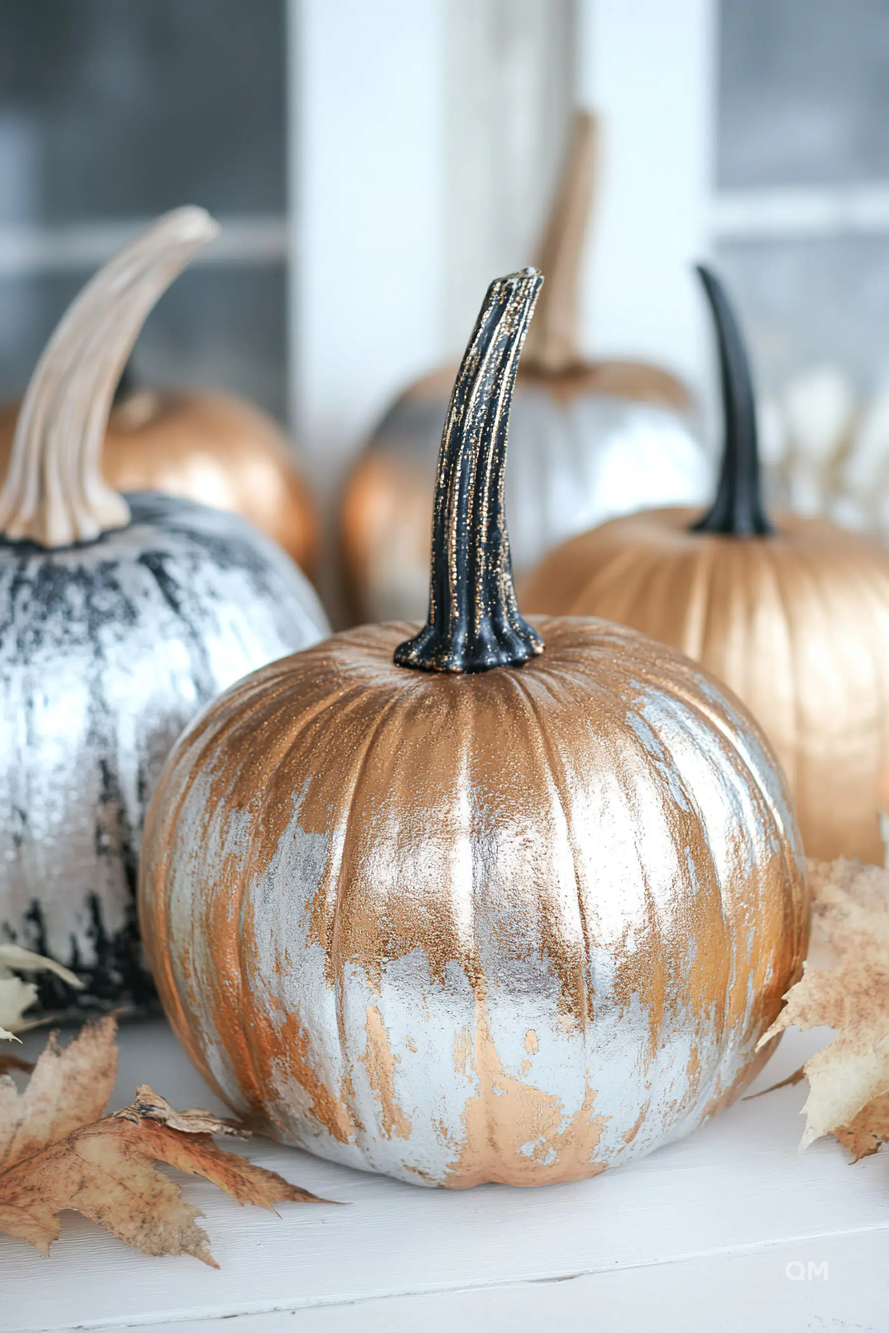 Decorative metallic-painted pumpkins in hues of gold and silver with dry leaves on a white surface.