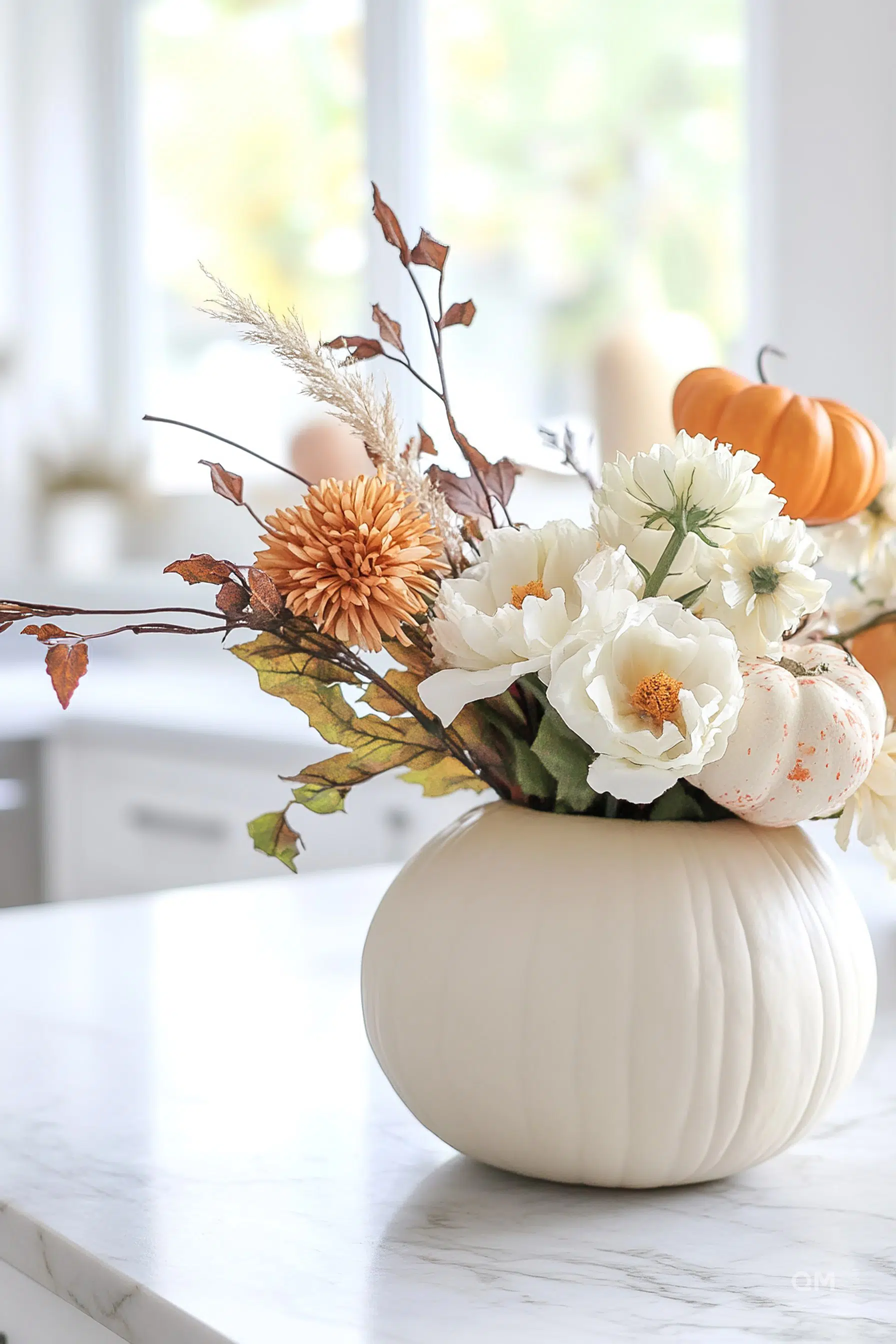 A white pumpkin vase on a marble countertop filled with autumnal flowers and leaves, with blurred window background and a small pumpkin.