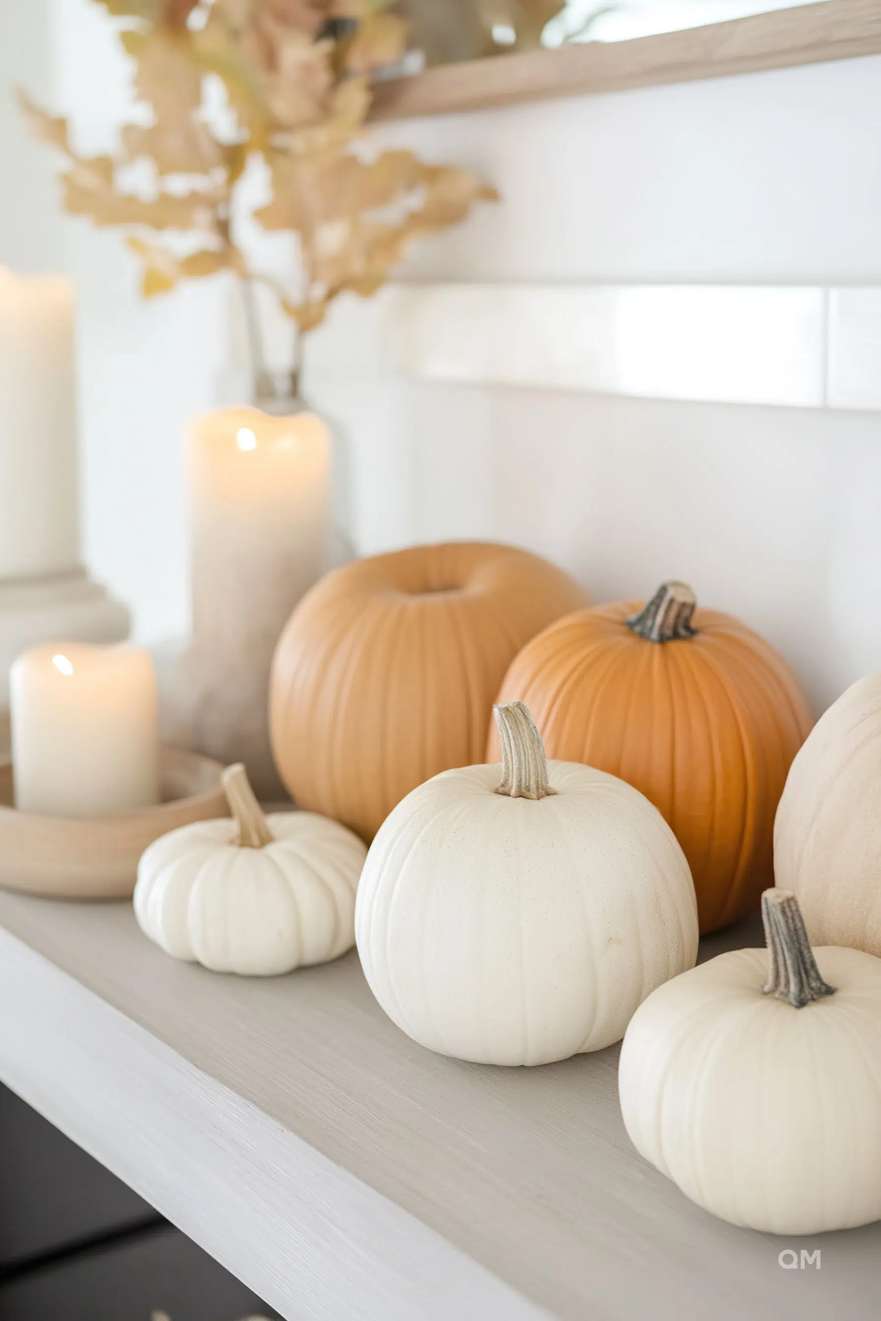 Decorative white and orange pumpkins on a shelf alongside lit candles and a vase with dried leaves, creating a calm autumnal vibe.