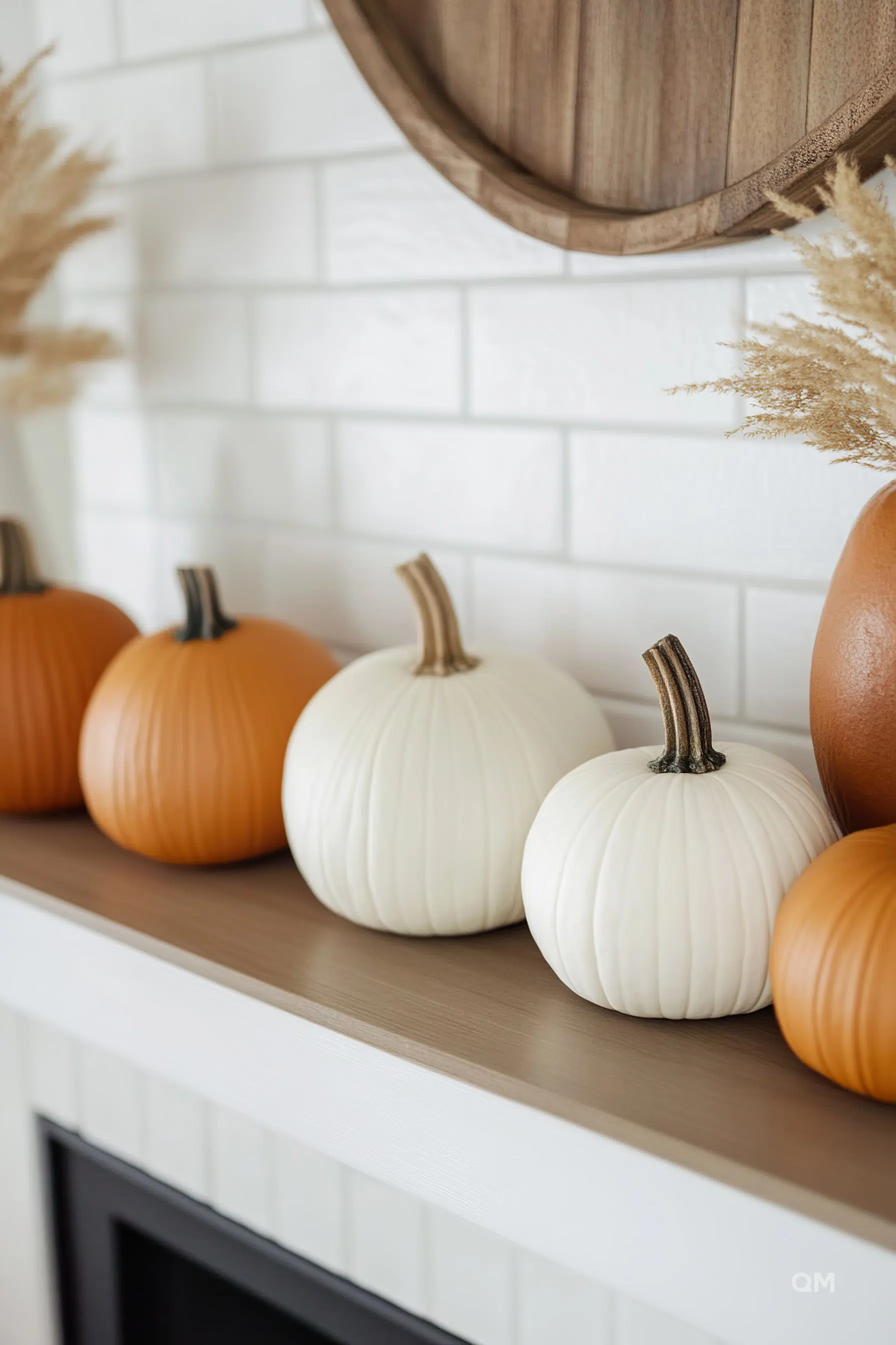 A row of decorative orange and white pumpkins on a mantelpiece with a white tiled background and dried pampas grass decoration.