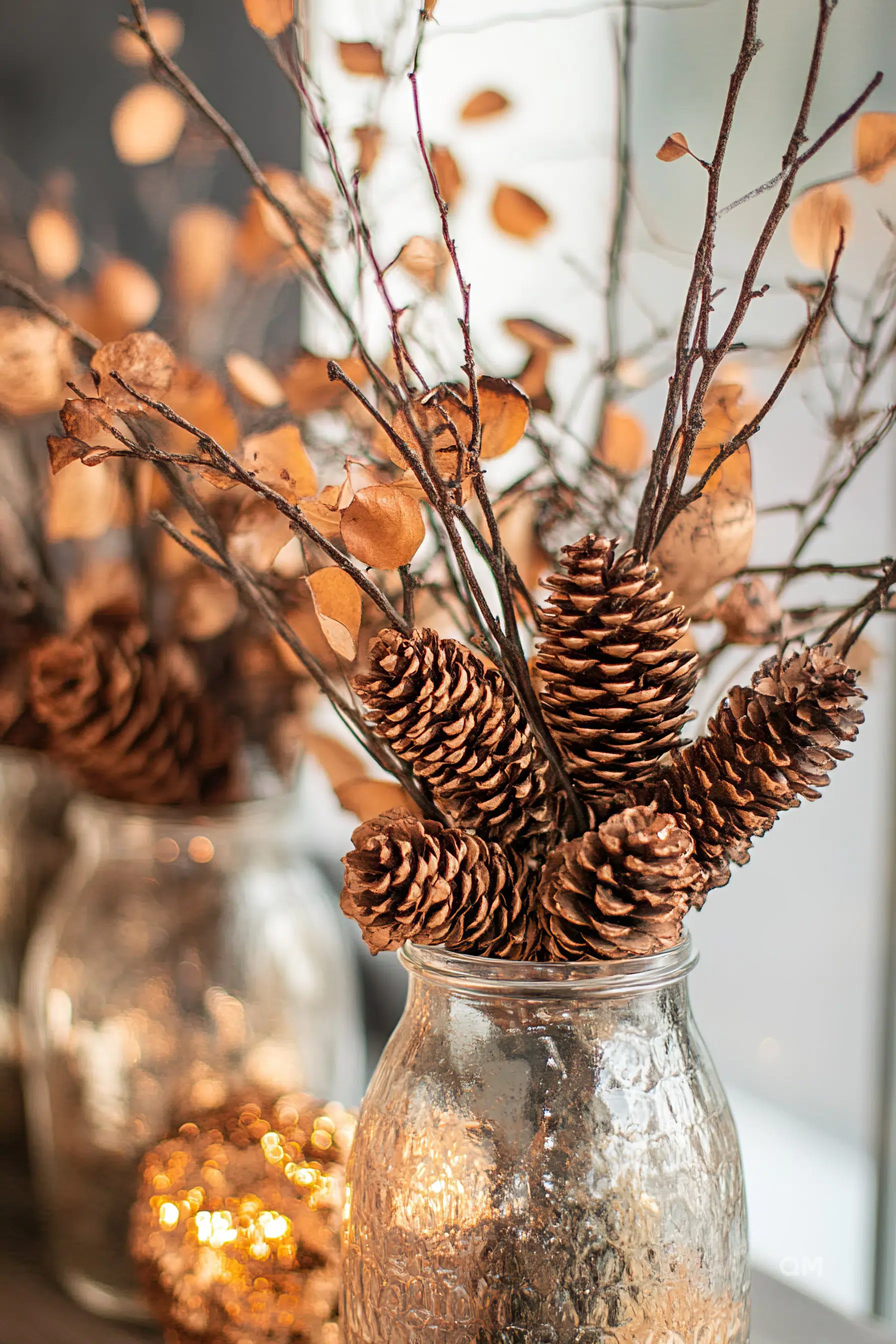 A rustic autumnal arrangement with pine cones and dried branches in a glass jar, highlighted by warm bokeh lights in the background.