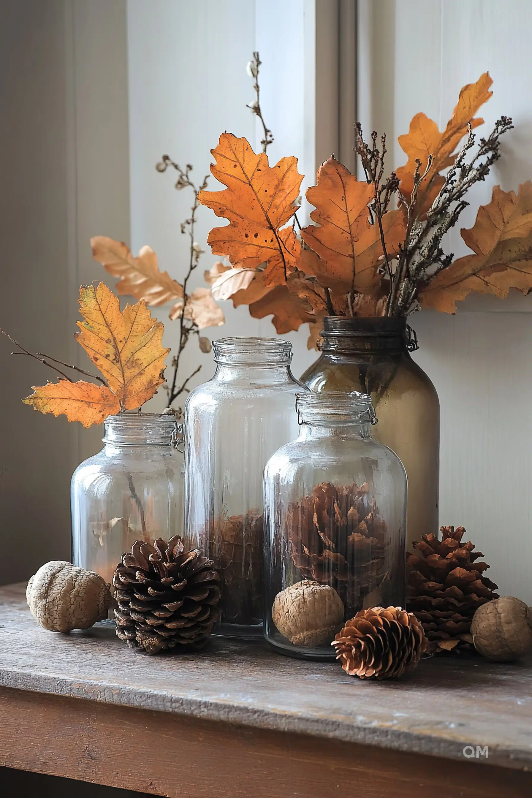 Alt text: A cozy autumnal display featuring glass jars with pine cones and dried leaves accompanied by scattered walnuts and pine cones on a wooden surface.
