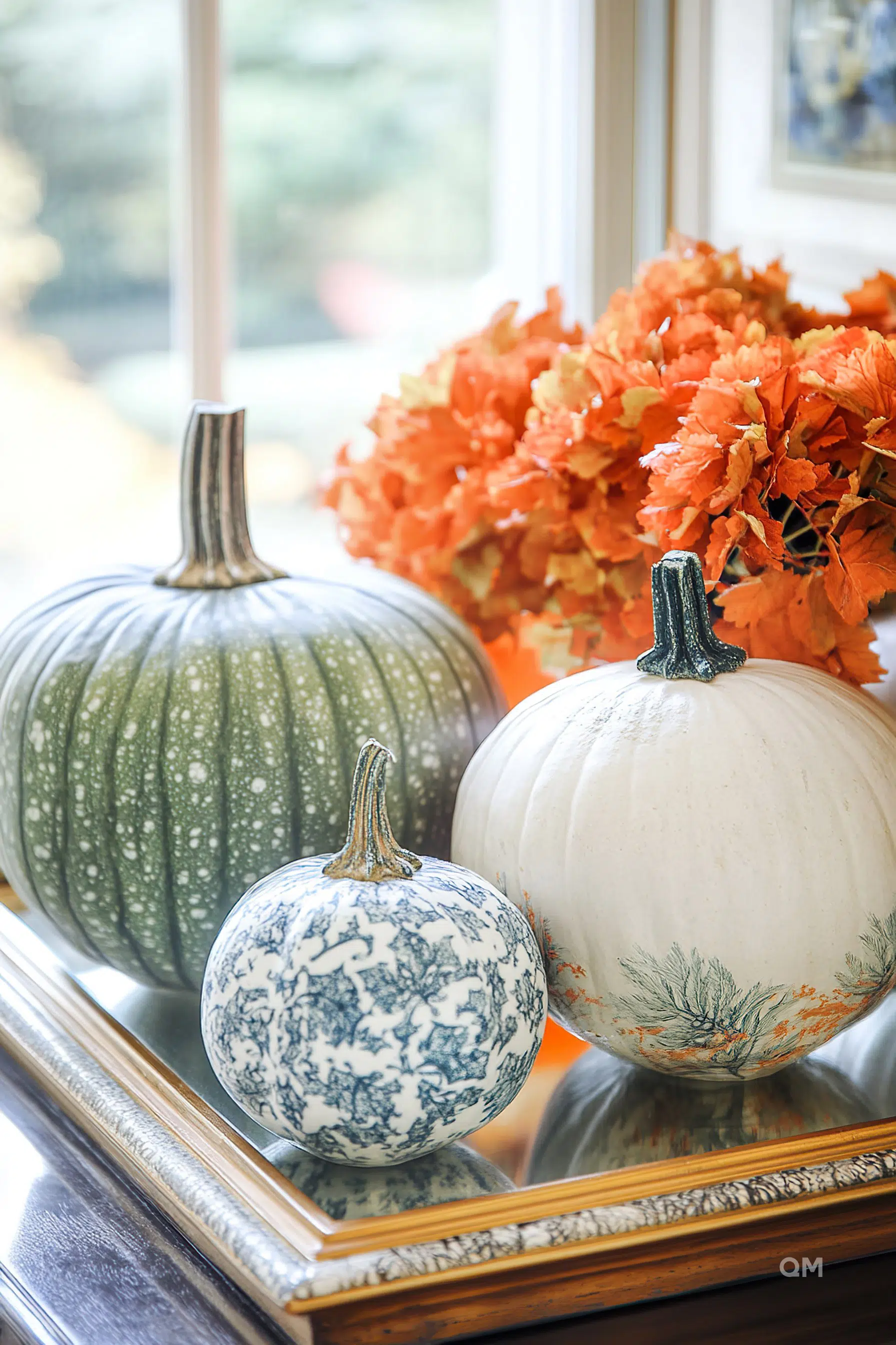 Three decorative pumpkins on a tray by a window, with patterns and colors suggesting autumn.