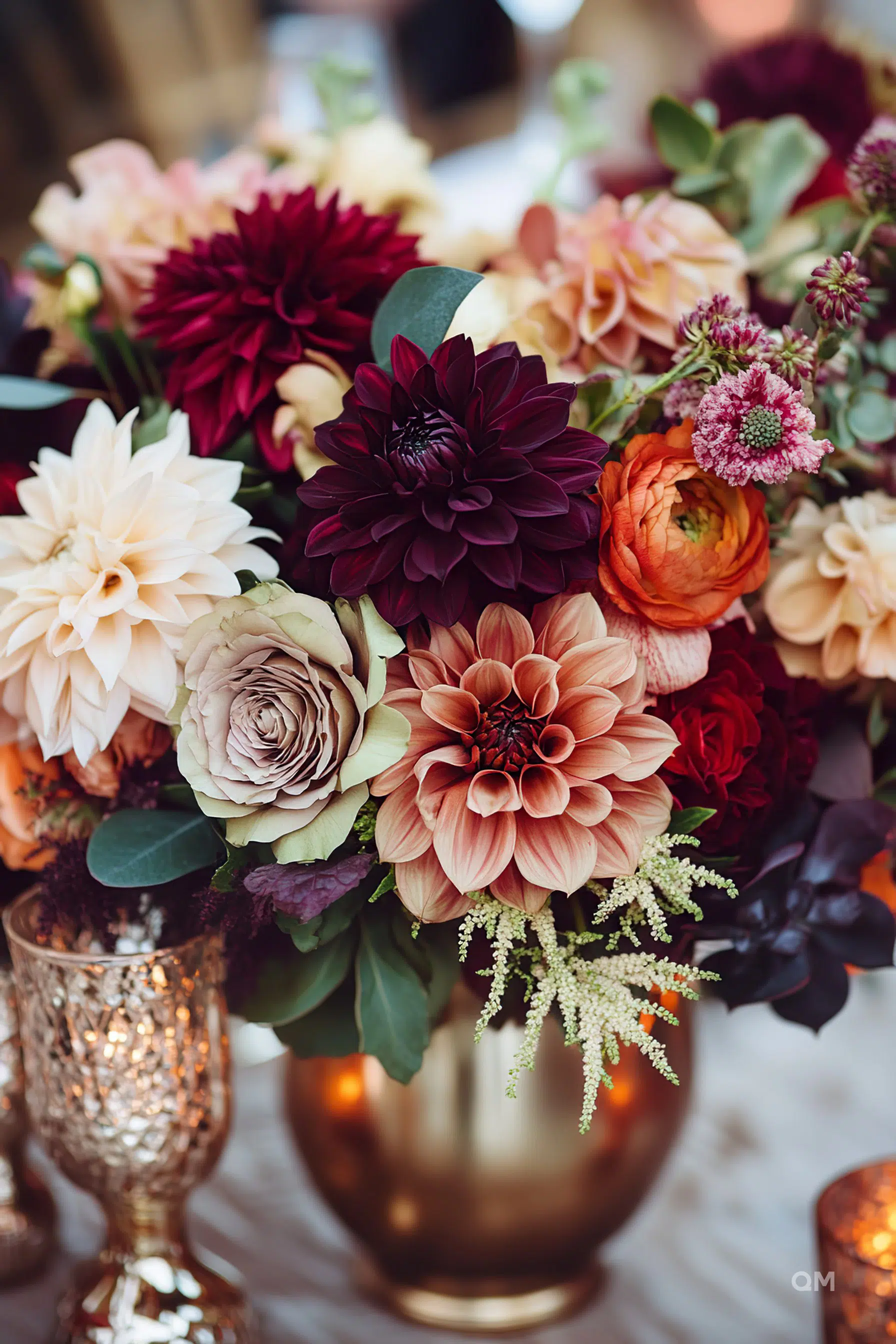Elegant bouquet with a variety of flowers including dahlias and roses in deep reds and pastel shades, arranged in a golden vase with subtle candlelight.